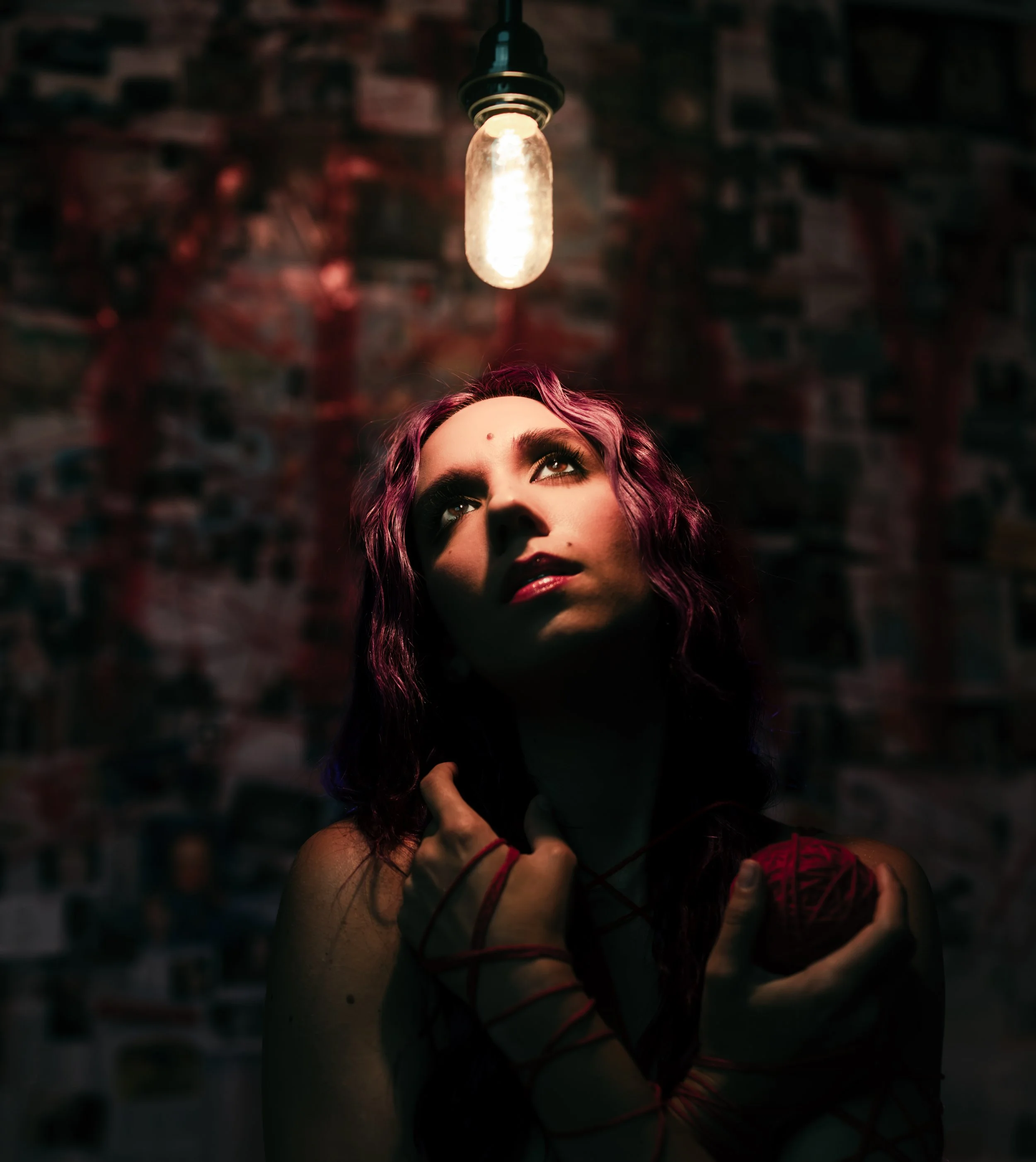Close-up of a woman's face with purple hair, looking upwards under a lit hanging bulb, against a blurred brick wall background.