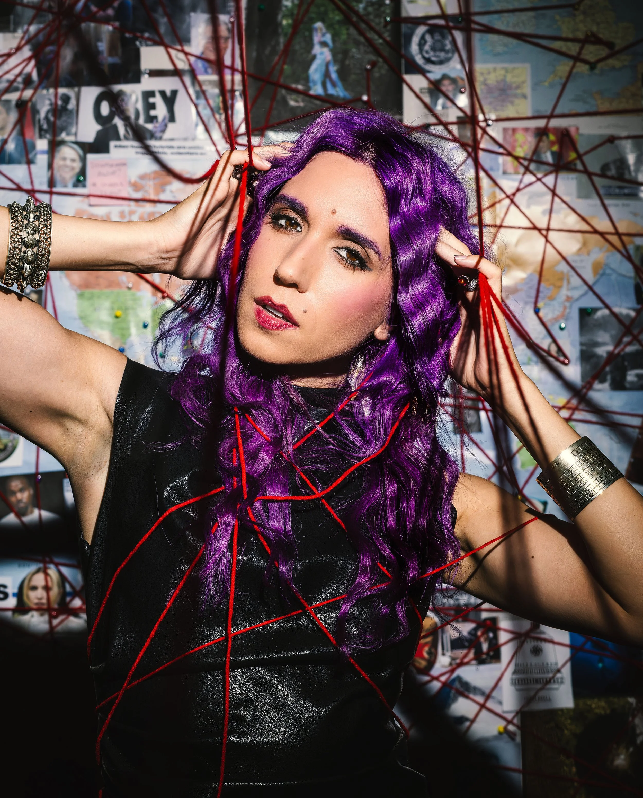 A woman with purple hair posing among red string art and a wall covered with photos, posters, and maps.