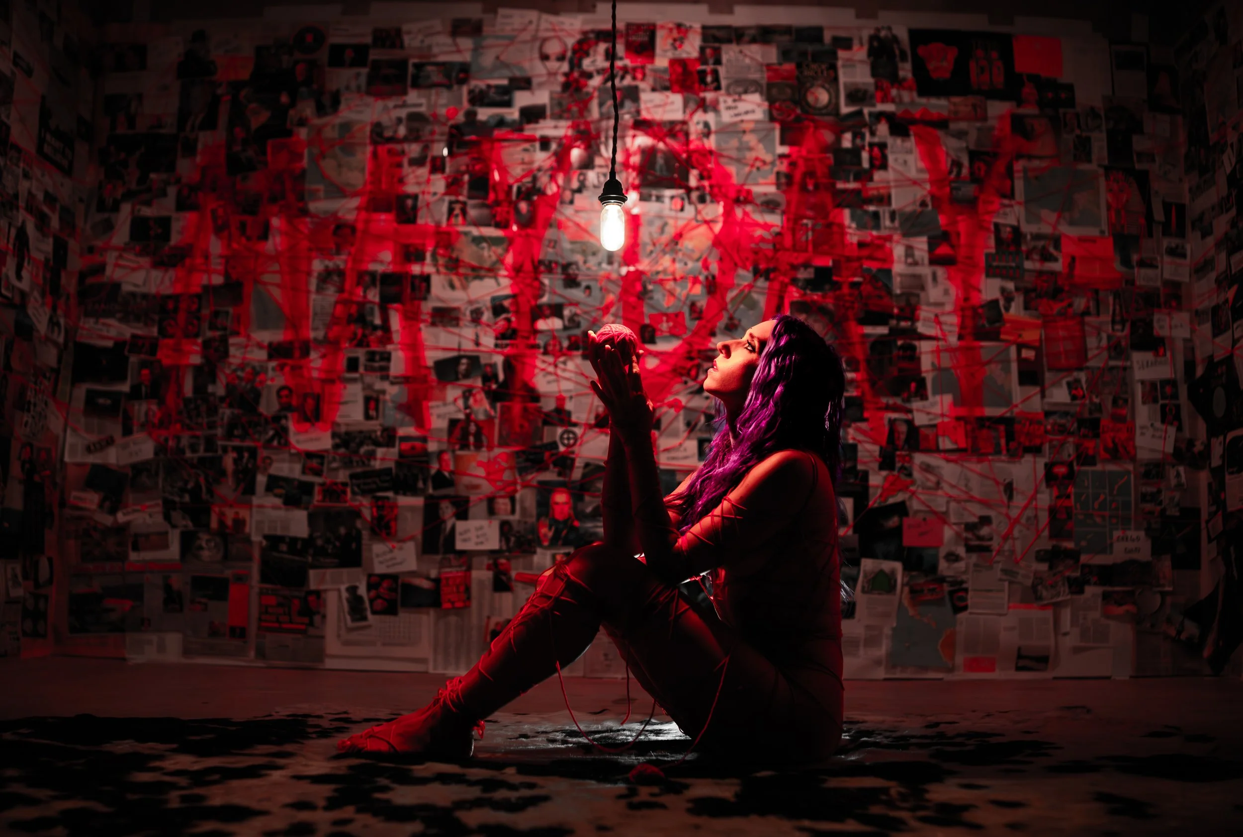 A woman with purple hair sitting on the floor with one knee raised, surrounded by red thread, in a dimly lit room with walls covered in photos and red markings.
