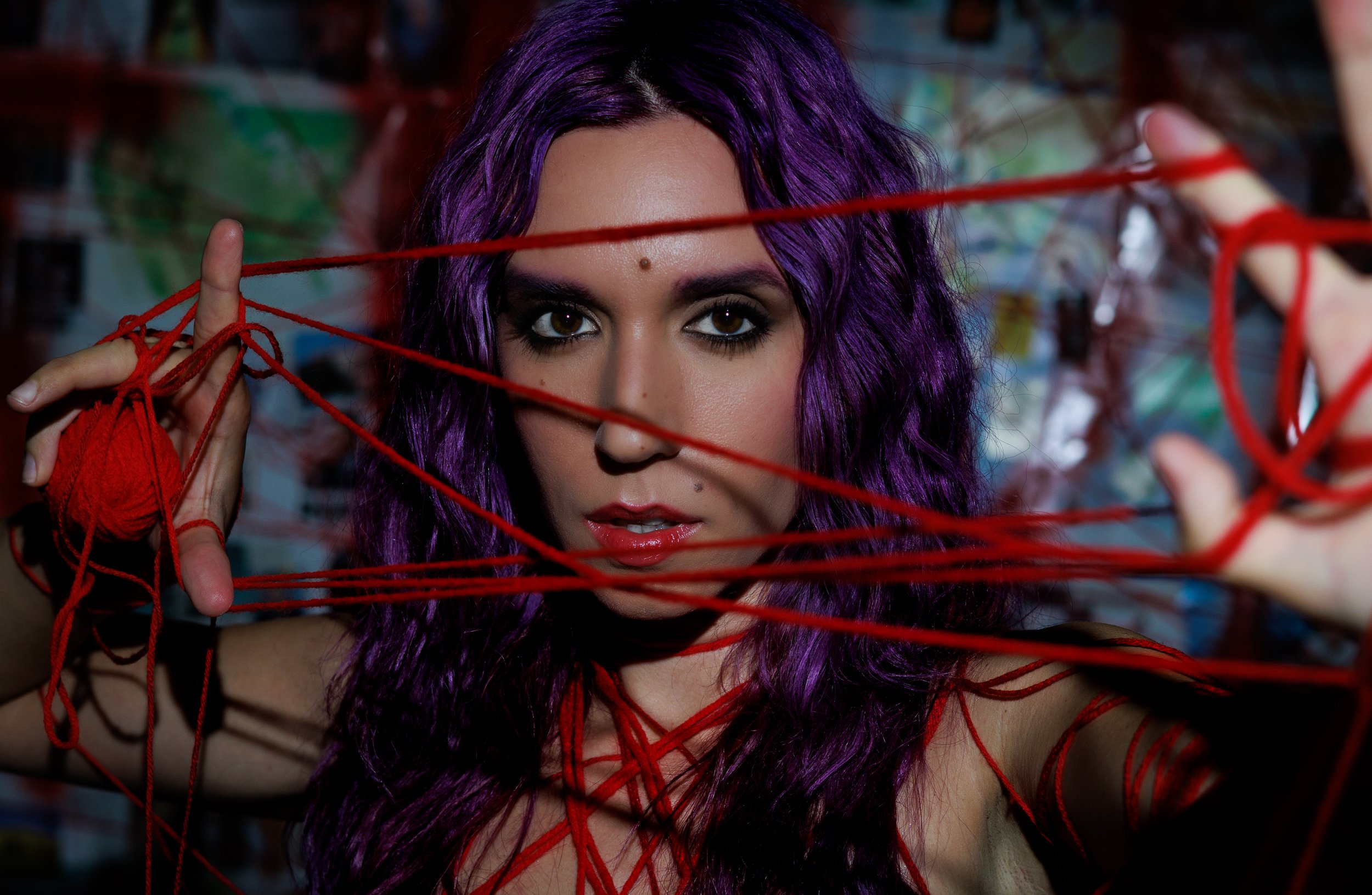 A woman with purple hair looking through red yarn strings held in her hands.