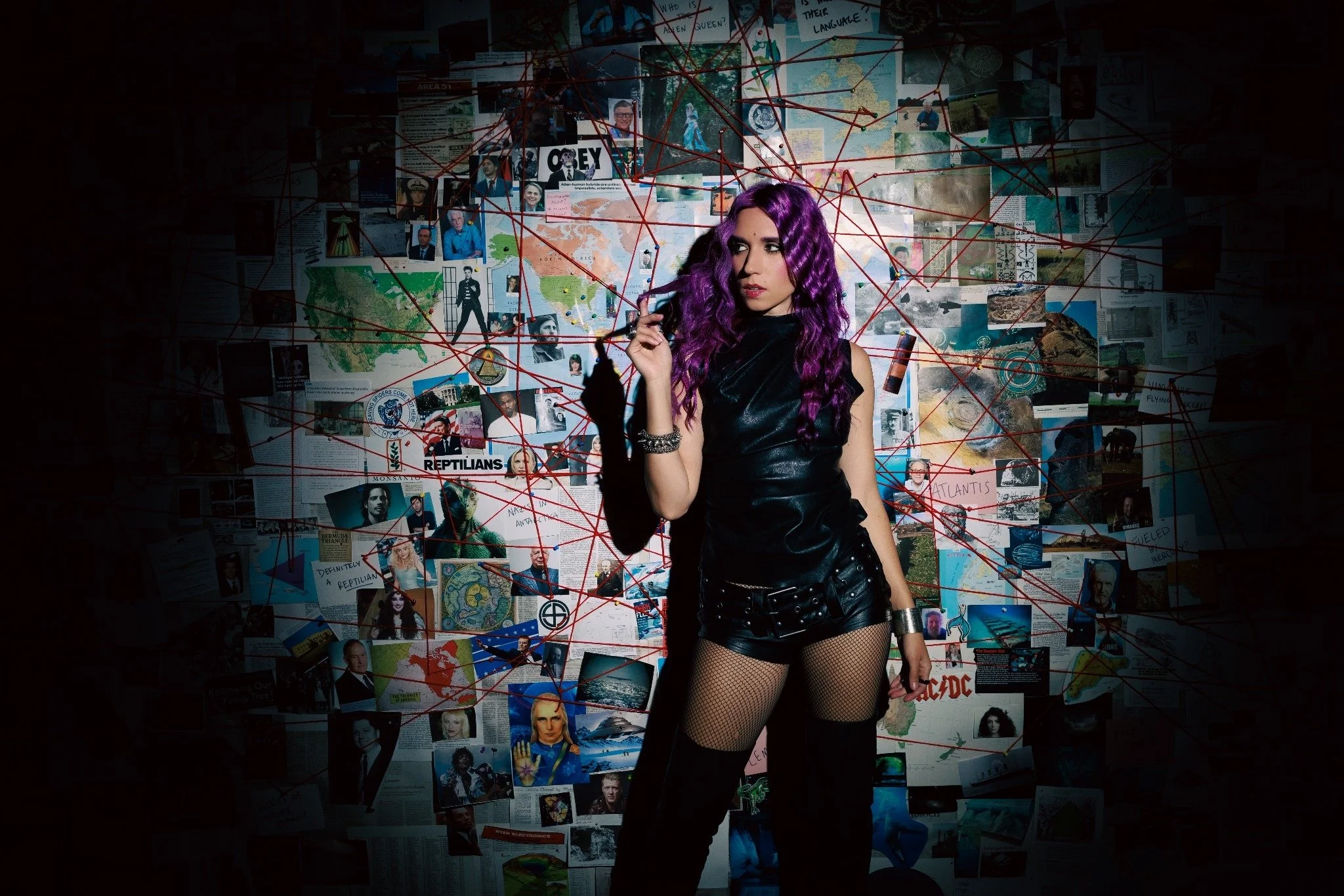 A woman with purple curly hair wearing a black leather dress, fishnet stockings, and black boots stands against a collage-covered wall with red string connecting various pictures and objects.