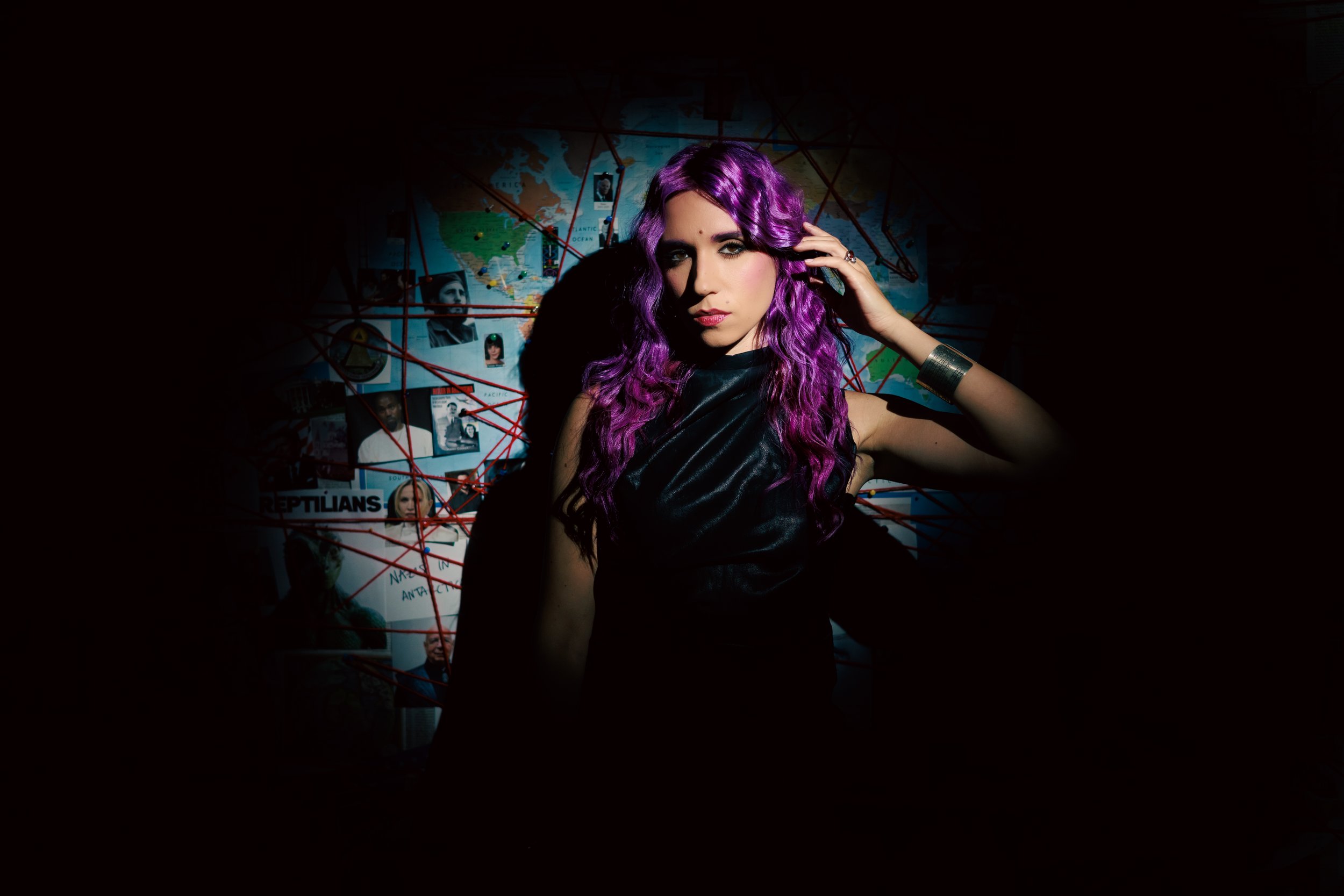 Young woman with long purple hair wearing black sleeveless top, touching her hair, standing in a dark room with a world map on the wall behind her, connected by red string to photos and notes.
