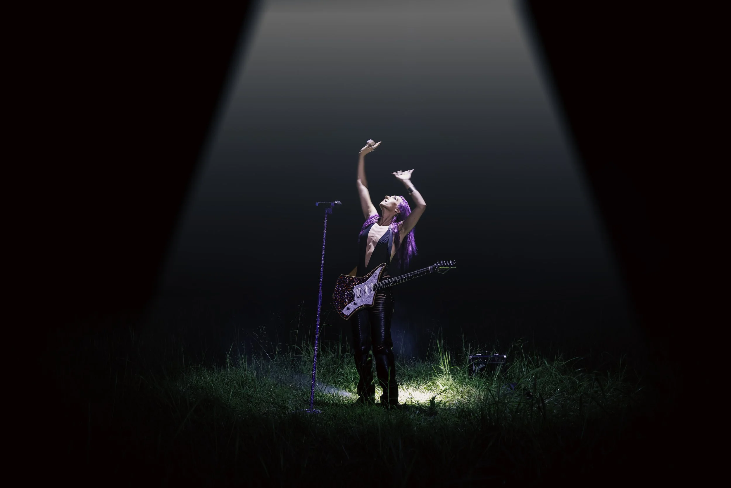 A female musician with colorful purple hair performing outdoors at night, standing in a spotlight with a guitar and a microphone on a grassy field.