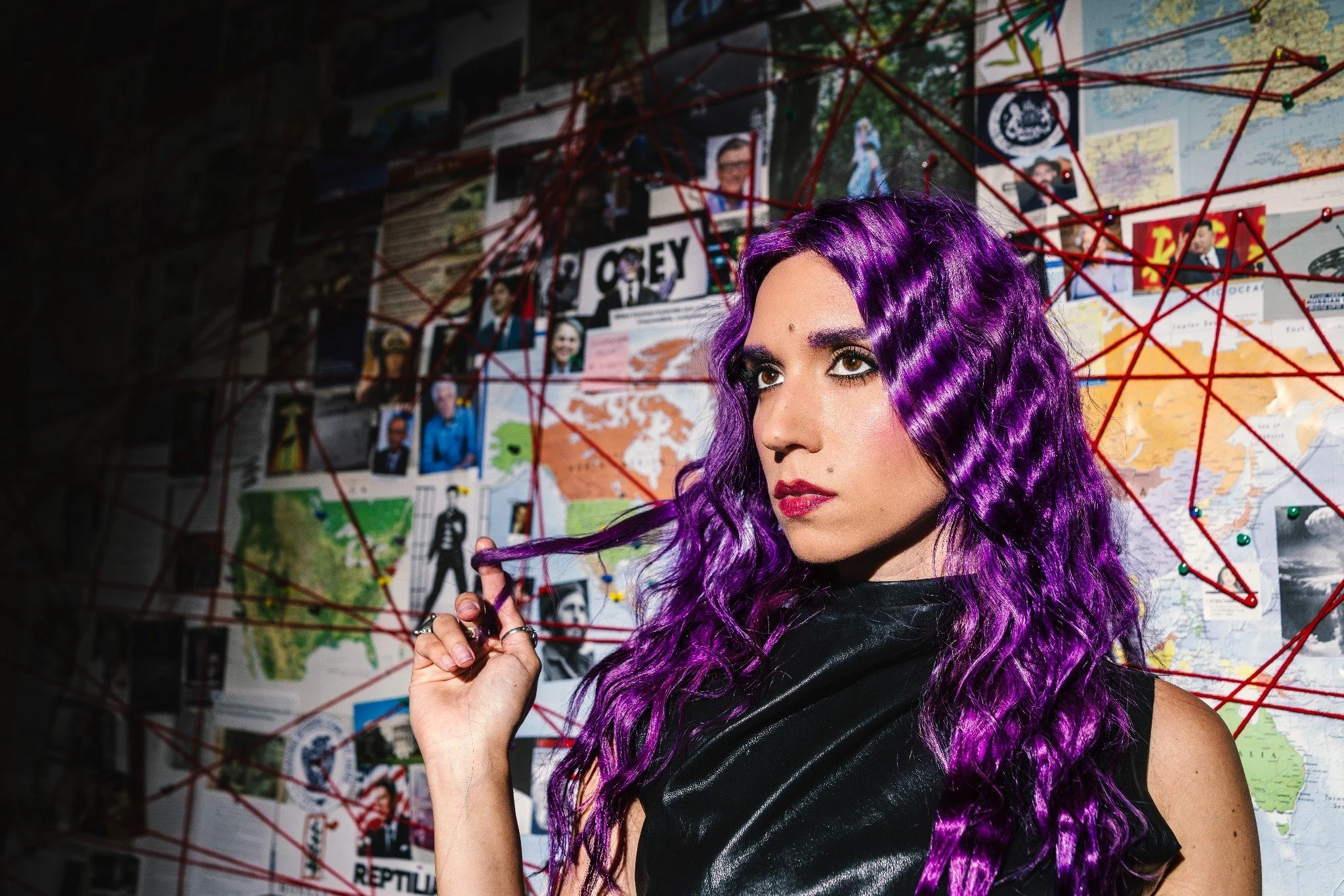 A woman with long, wavy purple hair and makeup, wearing a black outfit, stands in front of a wall covered with maps, posters, and red strings connecting various points.