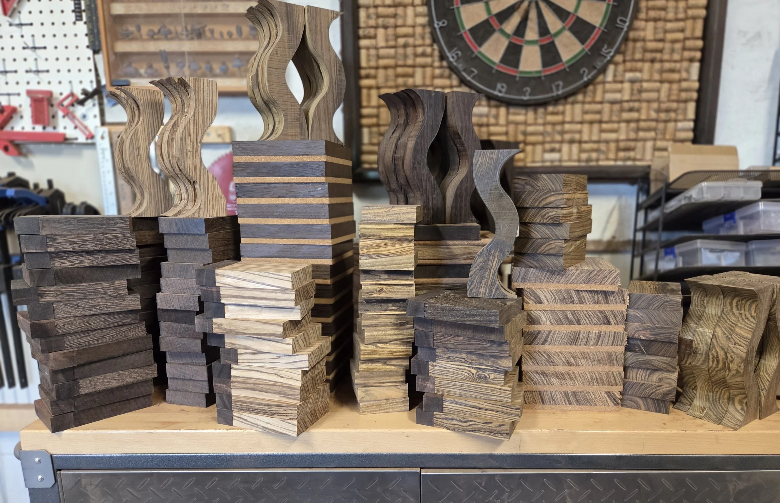 Stacks of unfinished wooden headphone stand components in various exotic hardwoods including wenge, Zebrawood, and bocote, arranged in a woodsroking stuio during early production stages for Grumpy Goose headphone Stands. A dartboard is visible.