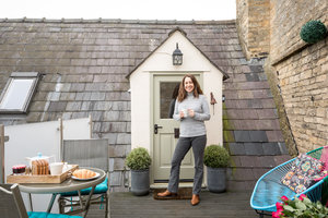 Amanda smiling while holding a mug outside the entrance of a cozy stone cottage, with a breakfast table and lounge chair set up nearby