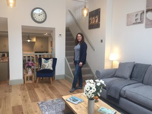 Amanda smiling in a stylishly furnished living room, complete with a comfortable blue sofa, a staircase, and home decor items like a clock and flowers