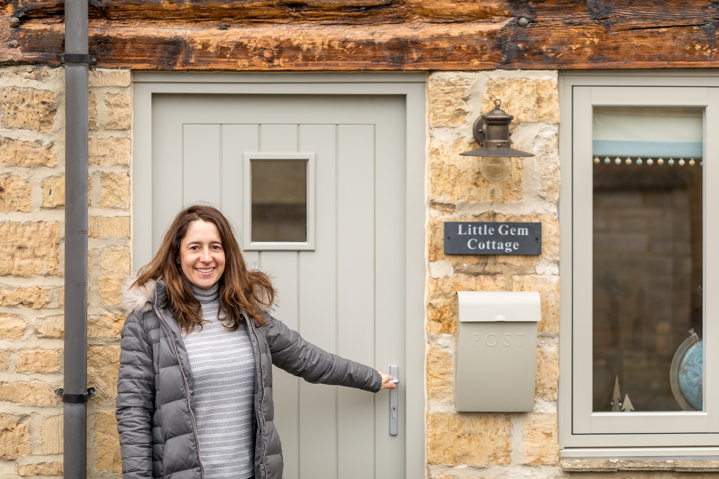 Amanda wearing a jacket stands outside the front door of Little Gem Cottage, gesturing toward the entrance