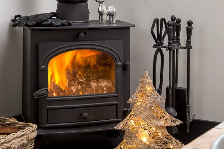 A close-up view of a cozy wood-burning stove with a  warm fire lit, featuring decorative items like a light-up tree figurine nearby
