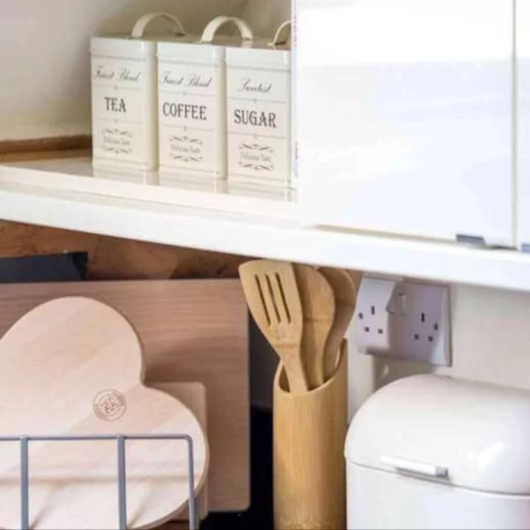 A close-up of a kitchen counter featuring labeled tea, coffee, and sugar canisters, alongside wooden kitchen utensils in a holder