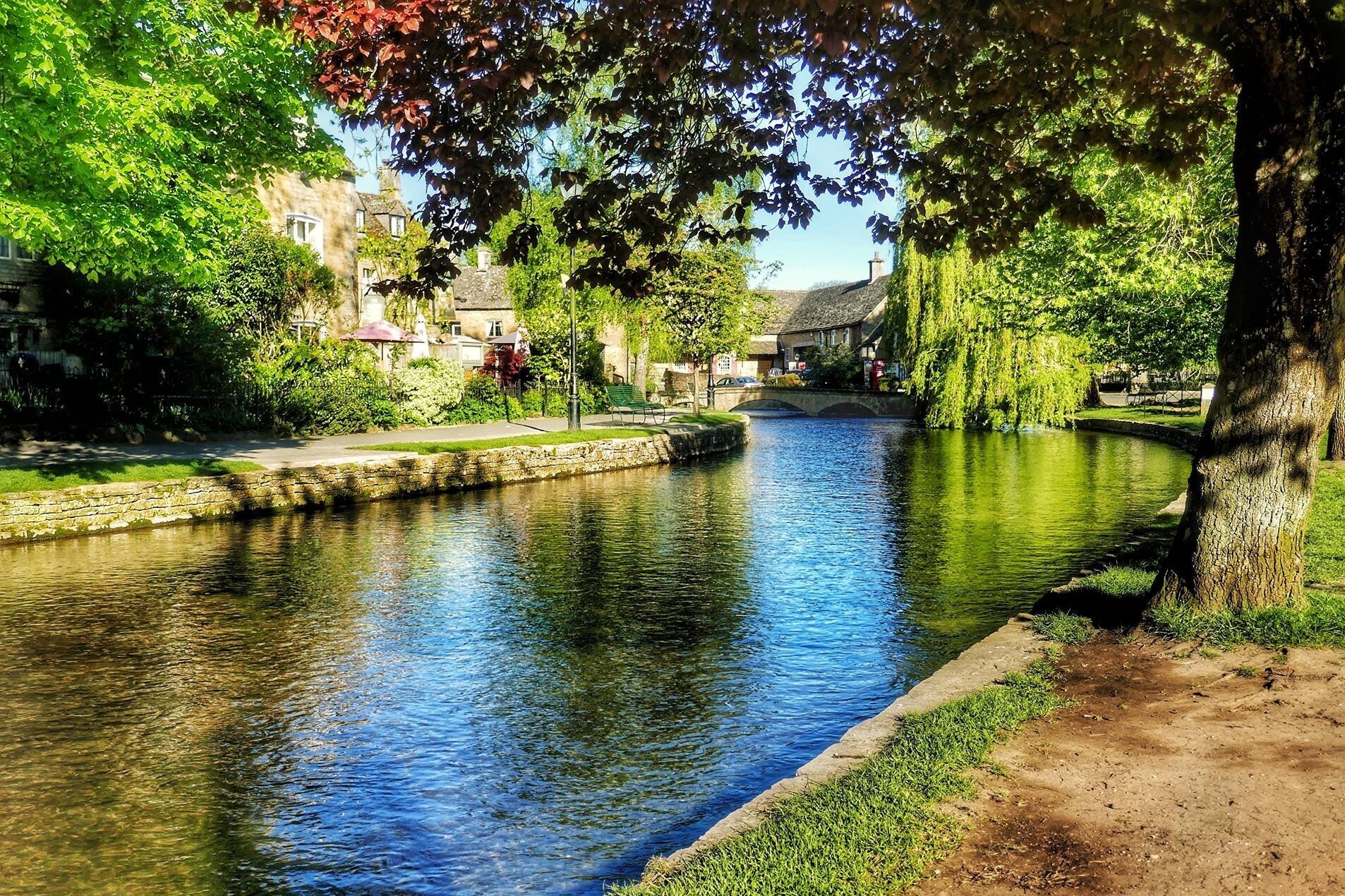 A picturesque view of the River Windrush flowing through Bourton-on-the-Water, surrounded by lush trees and stone buildings