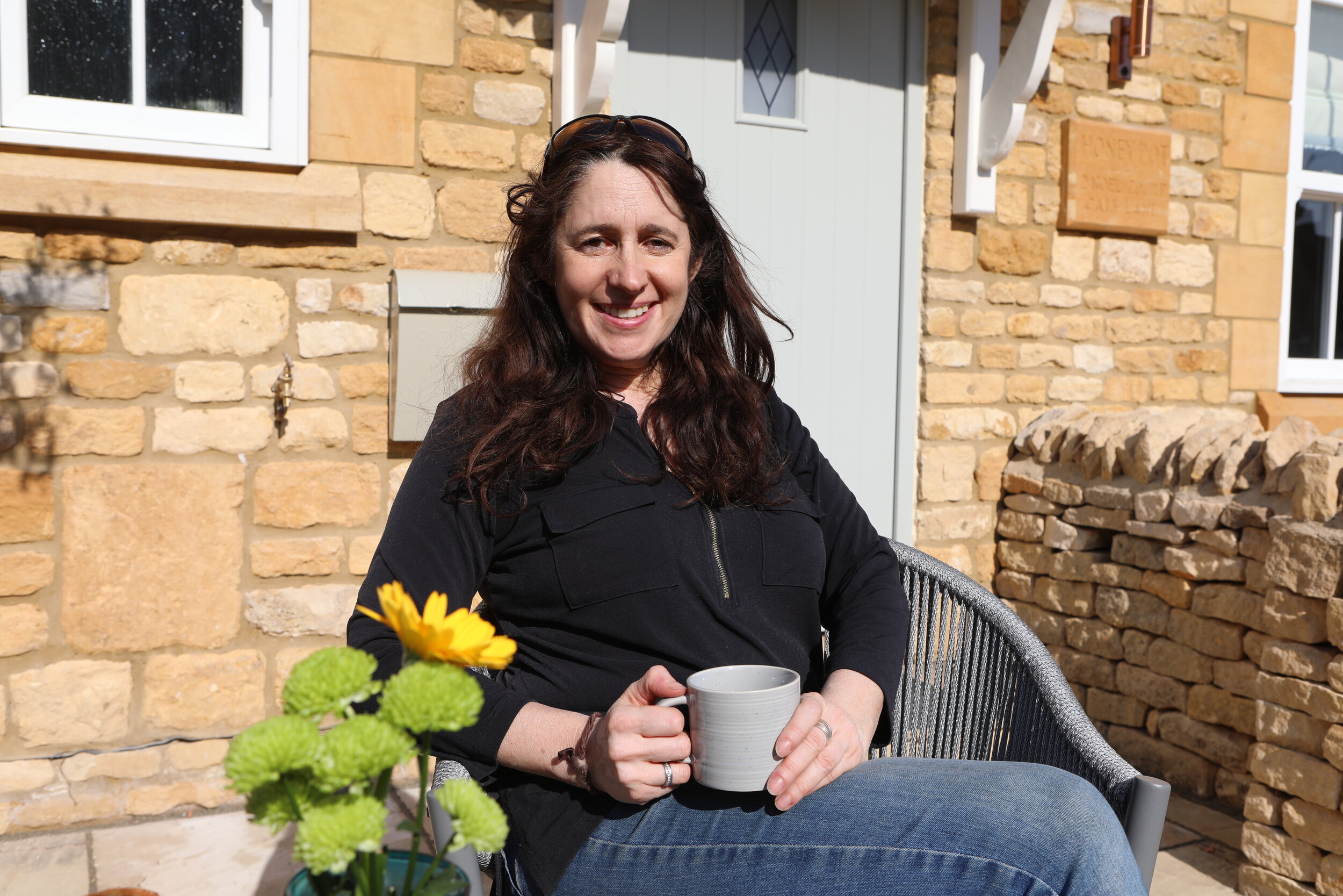 Amanda smiling identified as the owner of The Bolt Hole, sitting outdoors with a mug on a patio in front of a Cotswold stone wall