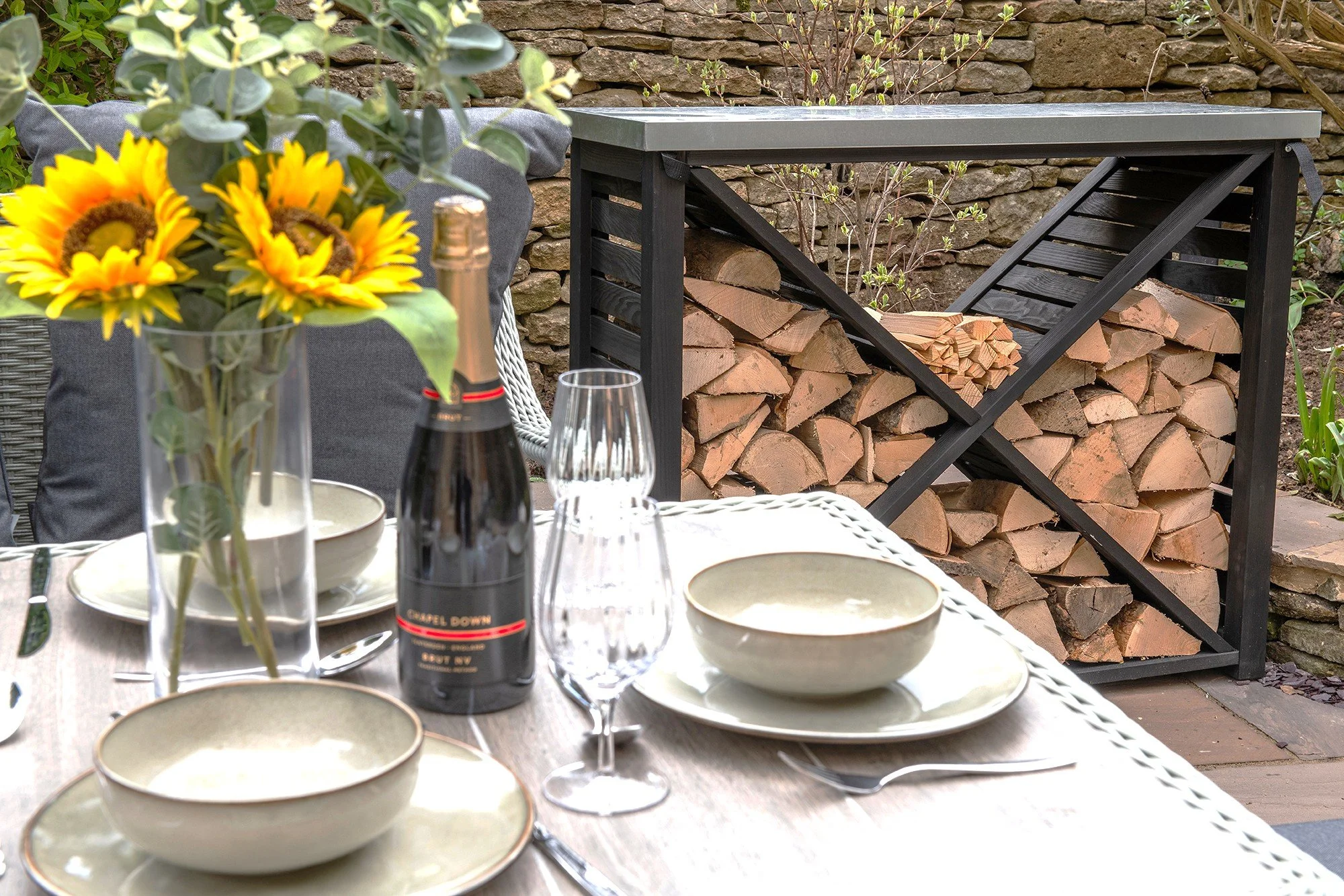 A charming outdoor dining setup in a Cotswolds cottage garden featuring a vase of sunflowers, a bottle of sparkling wine, and a neatly stacked log store in the background