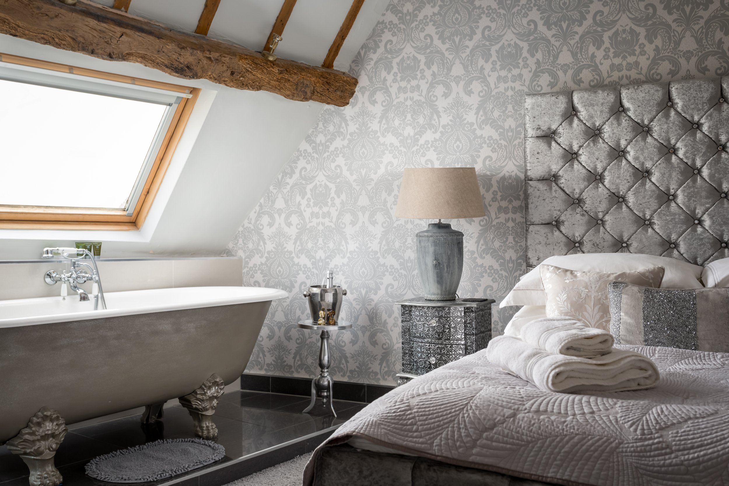 A luxurious, spacious bedroom at The Loft in Stow-on-the-Wold, featuring a  roll-top bath, elegant patterned wallpaper, and a cozy bed
