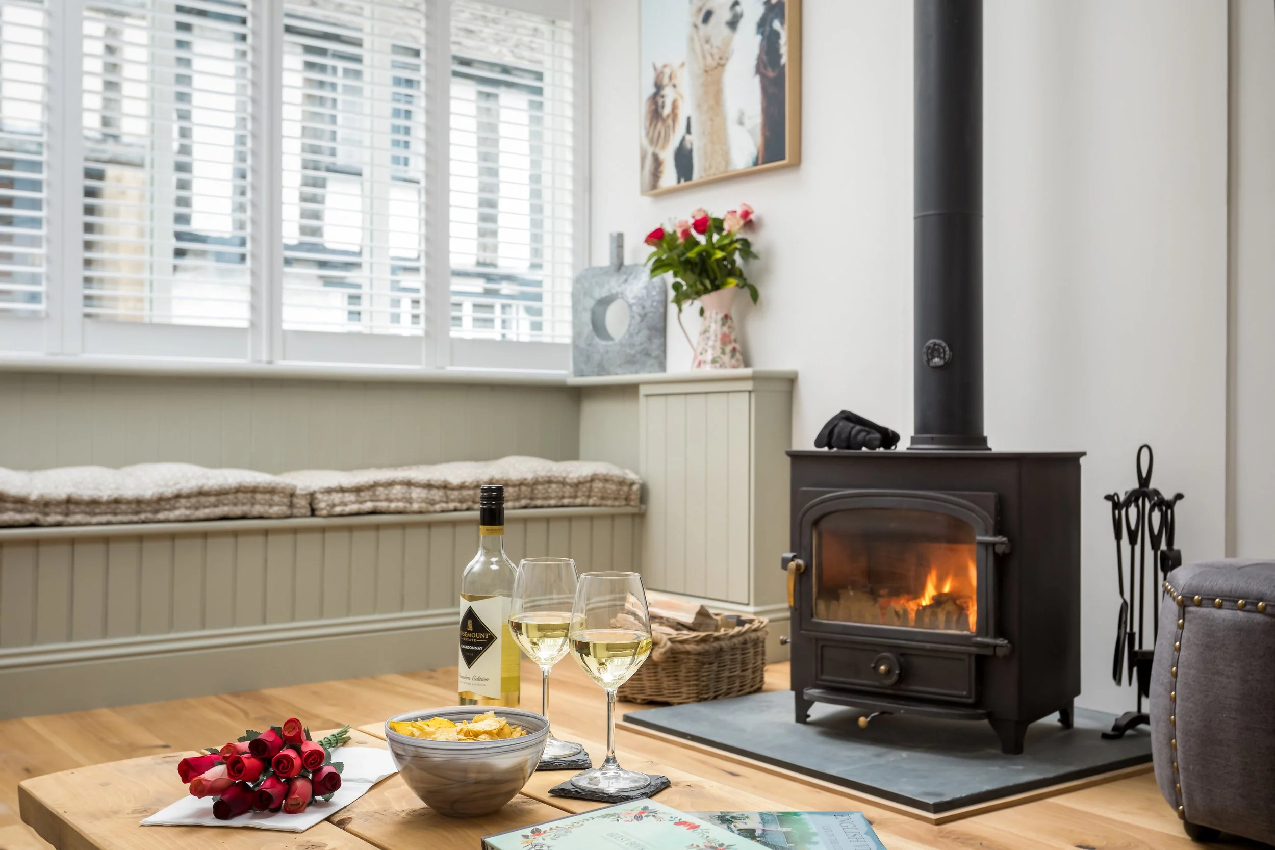 A cosy and inviting living are at The Bolt Hole featuring a lit wood-burning stove, glasses of wine, and a bowl of grapes, creating a relaxing holiday atmosphere