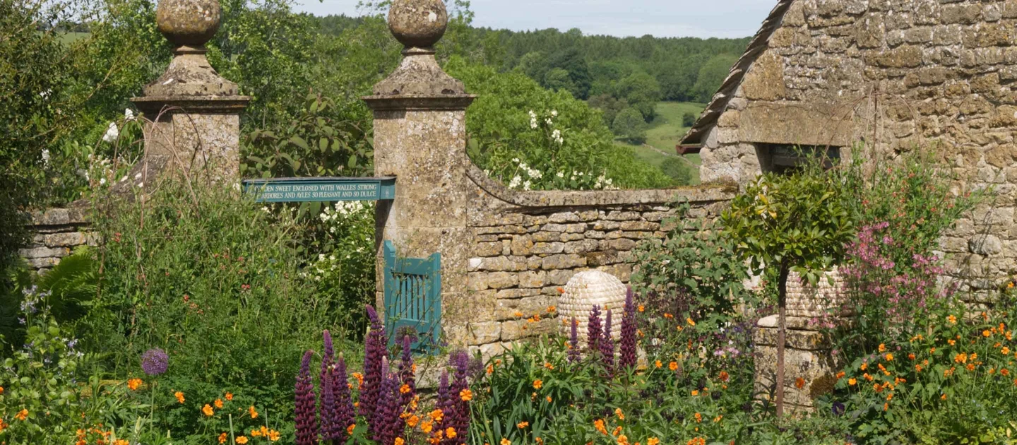 Scenic view of the terraced hillside garden at Snowshill Manor, featuring historic stone walls, large stone pillars framing a green gate, and a variety of blooming purple and orange flowers