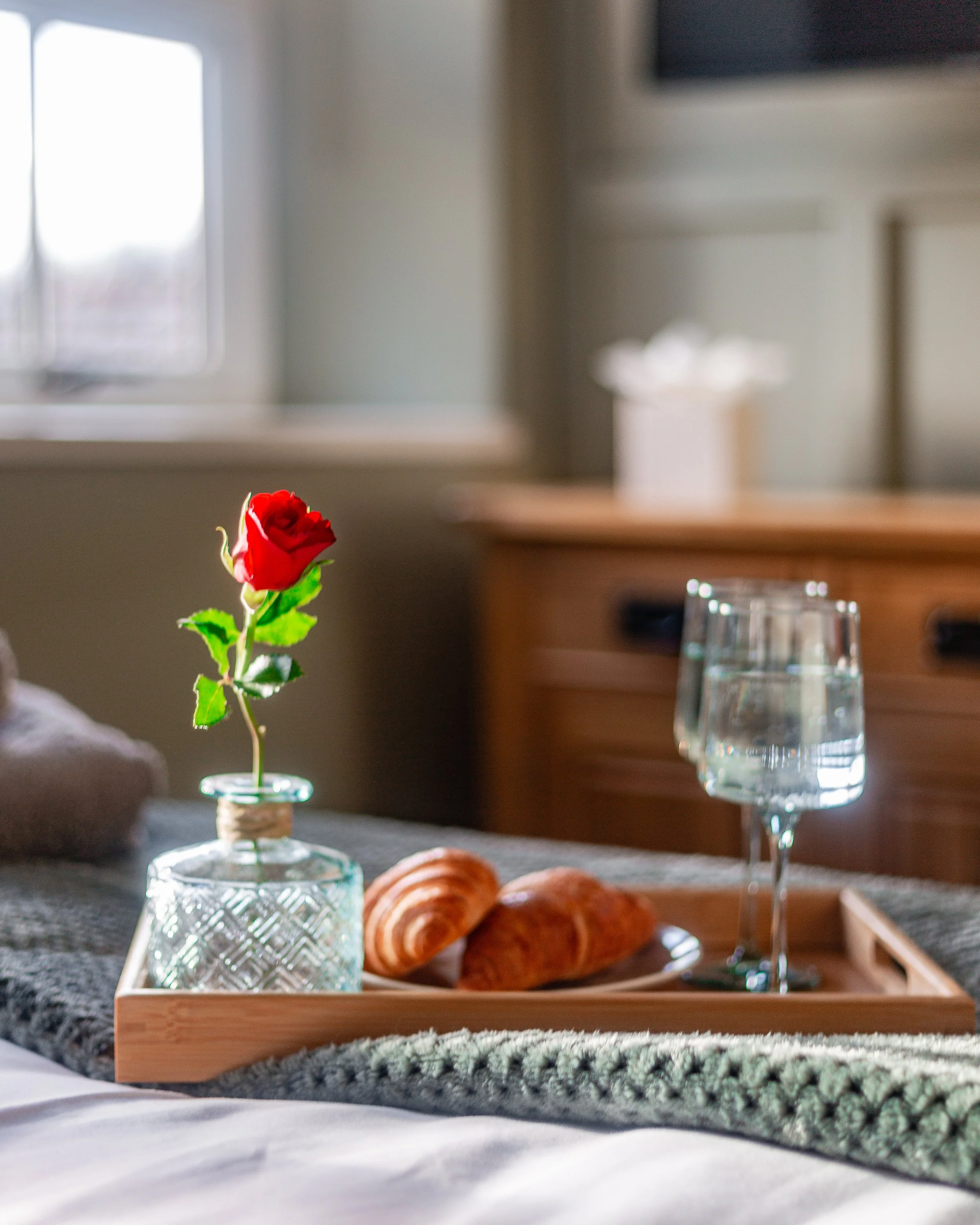A cozy breakfast tray set on a bed, featuring a single red rose in a glass vase.