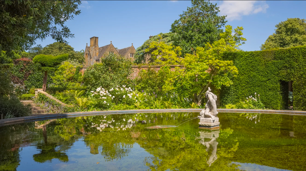 A tranquil pond reflecting the surrounding lush greenery and architecture at Hidcote Manor Garden, one of England's most celebrated Arts and Crafts gardens