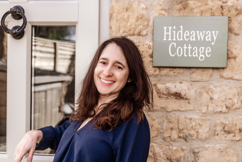 A smiling portrait of Amanda Stecker, the owner of Hideaway Cottage, set against a traditional Cotswold stone wall with the cottage's nameplate visible, located in Bourton-on-the-Water
