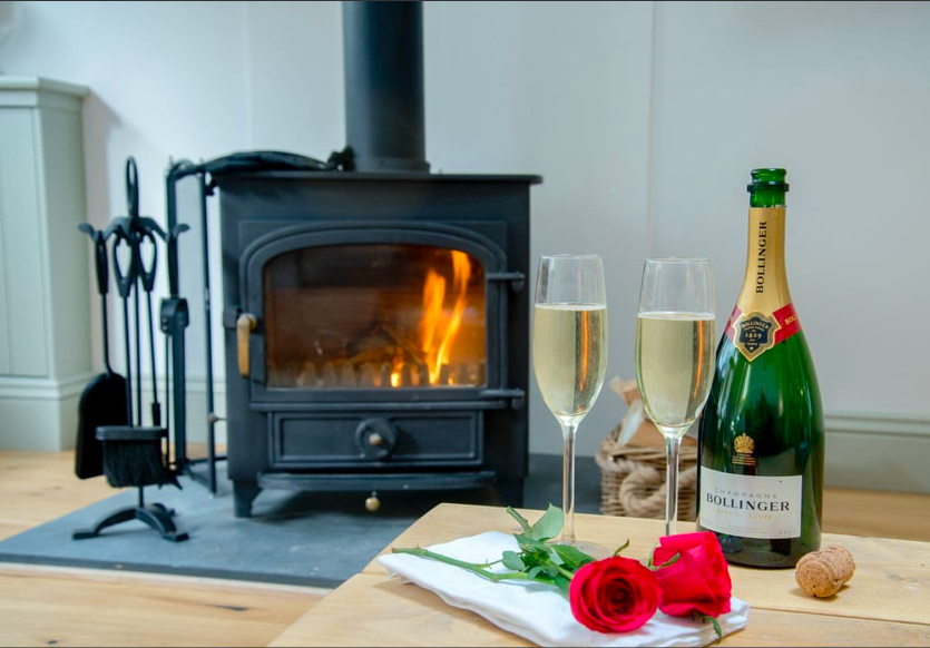 A cosy setting at The Bolt Hole in Winchcombe featuring a warm log burner, two glasses of champagne, and a bottle, creating a romantic atmosphere