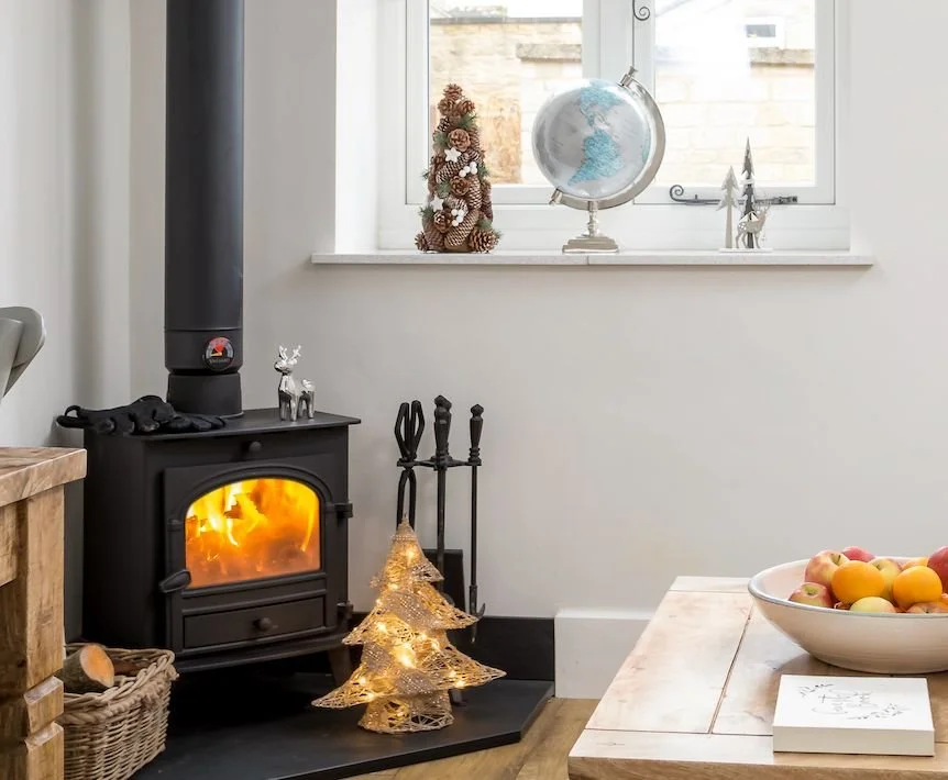 A cosy cottage interior featuring a burning wood stove, festive Christmas decorations including a light-up tree, and a bowl of fruit on a wooden table