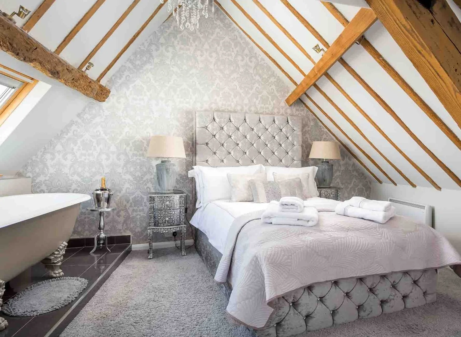 A luxurious, spacious bedroom in The Loft apartment, featuring vaulted ceilings with exposed beams, a plush bed, and a stylish roll-top bath