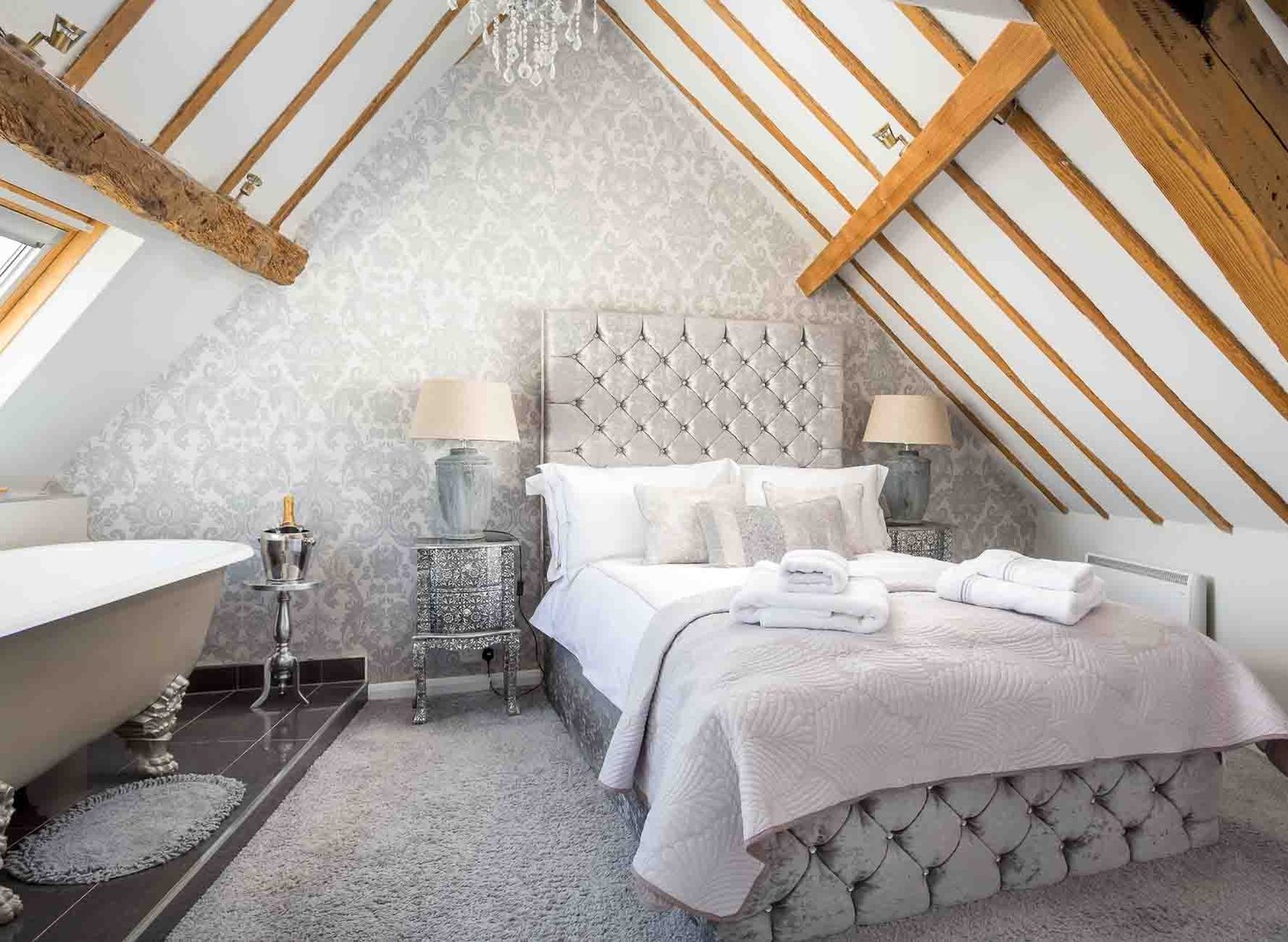 Luxury attic bedroom at The Loft in Stow-on-the-Wold featuring an upholstered bed, patterned feature wall, exposed wooden roof beams, and a freestanding roll-top bath on a raised tiled platform