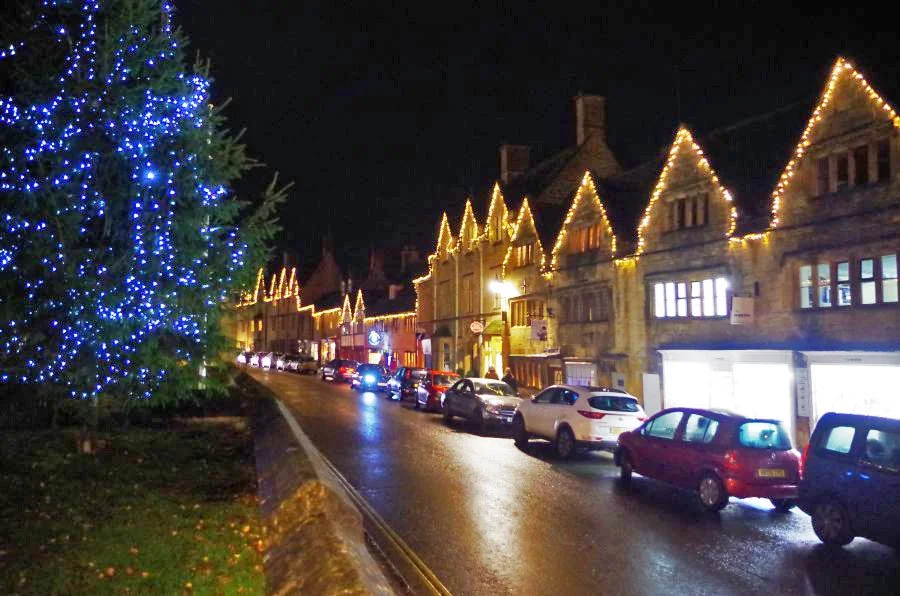 A festive evening scene in Chipping Campden featuring illuminated buildings and Christmas lights along the street