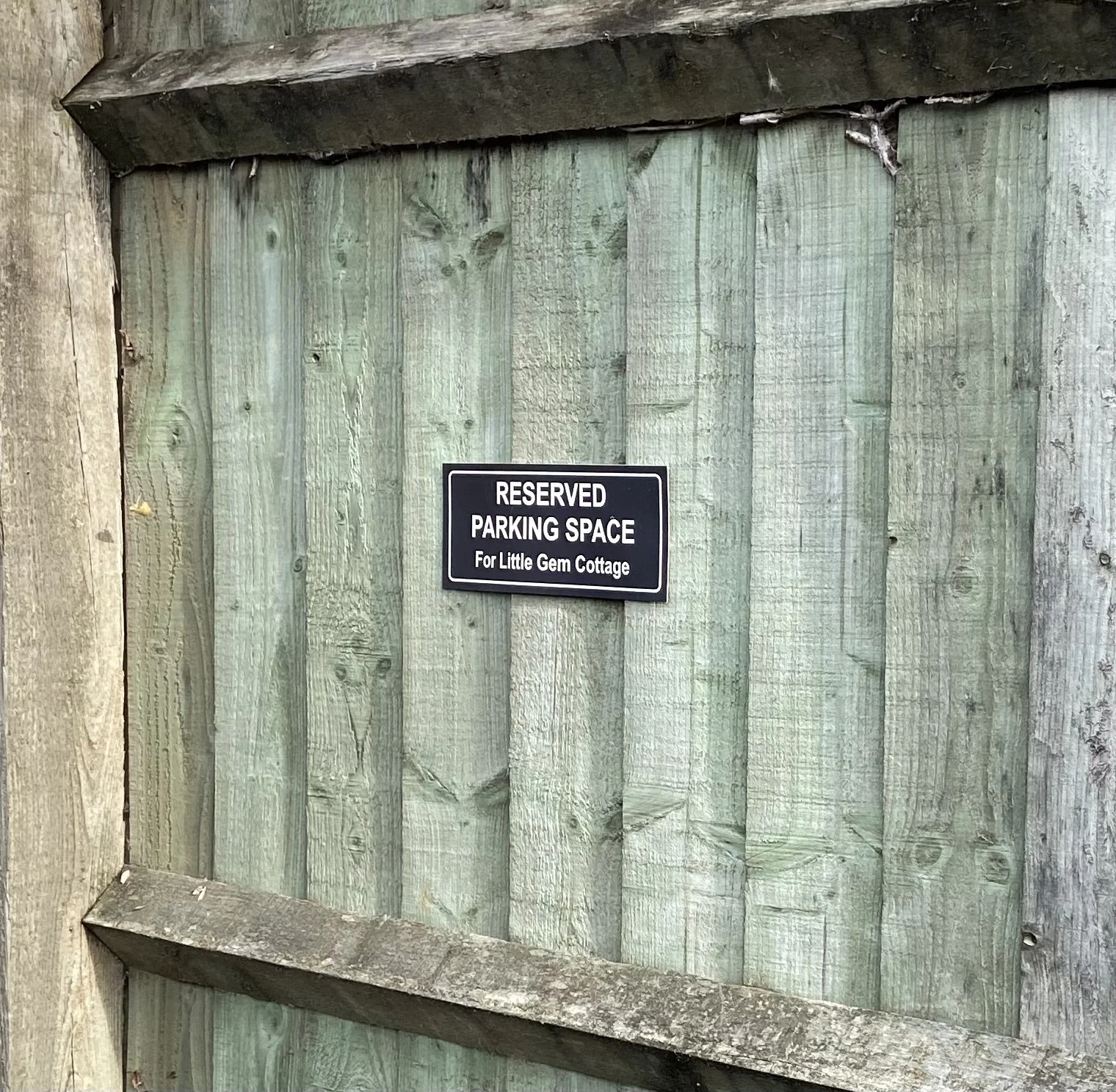 Close-up view of the weathered grey wooden fence at Little Gem Cottage in Bourton-on-the-Water, featuring a black rectangular sign that reads 'RESERVED PARKING SPACE For Little Gem Cottage'
