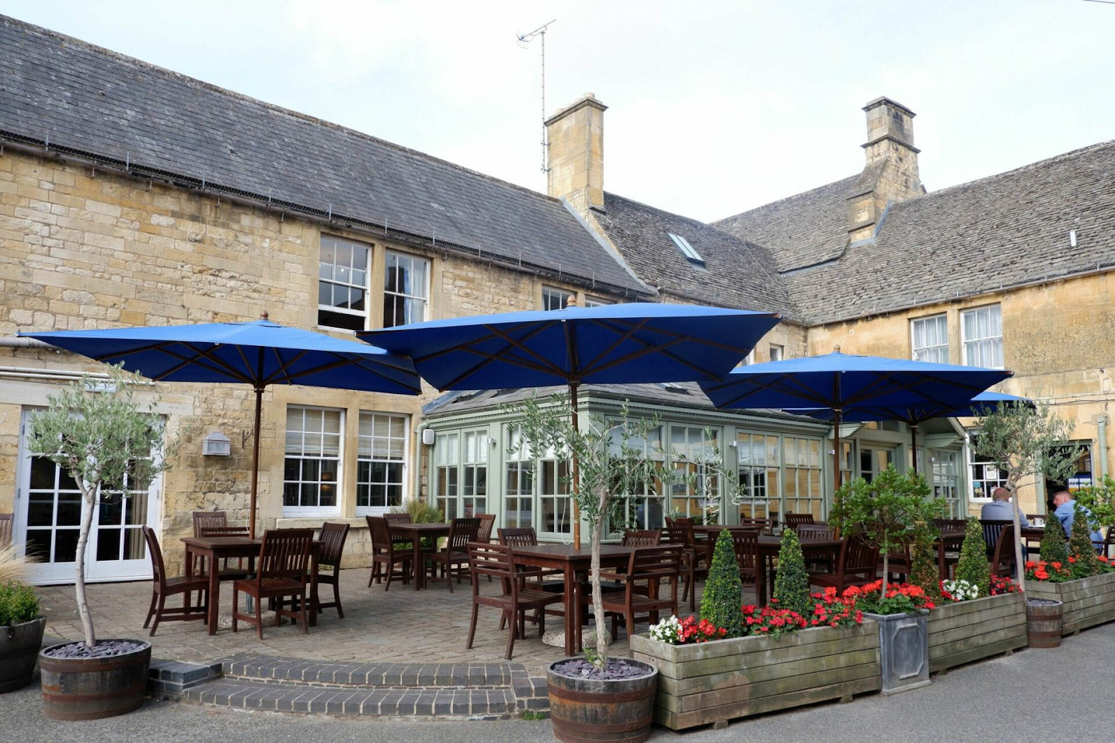 Outdoor patio and dining courtyard at The Noel Arms pub in Chipping Campden, featuring wooden tables and chairs under large blue umbrellas next to a historic stone building