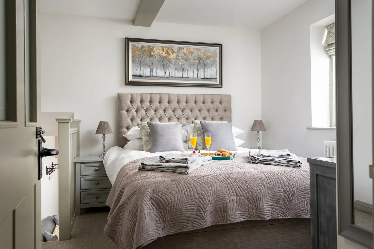 Boutique bedroom at The Bolt Hole cottage in Winchcombe featuring a king-size bed with a large tufted headboard, a breakfast tray with orange juice on the bed, and a framed forest picture on the wall