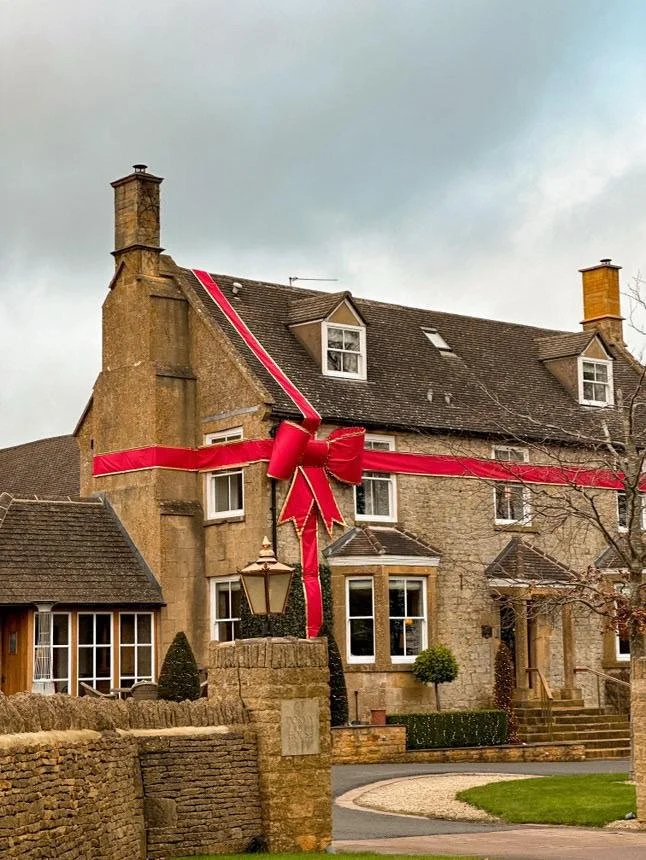 A traditional stone cottage in the Cotswolds wrapped with a large red ribbon and bow, representing a holiday getaway gift