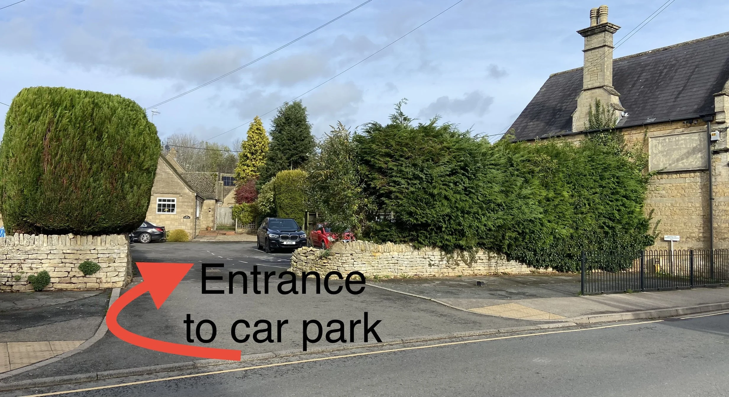 External view of the stone wall entrance leading to the parking area for Little Gem Cottage in Bourton-on-the-Water, featuring a large red arrow pointing left with text overlay reading 'Entrance to car park'