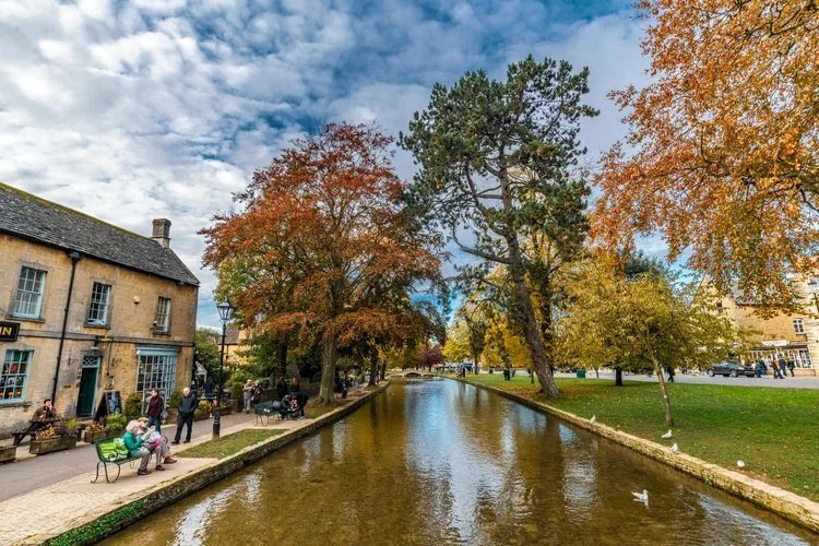 A scenic view of the River Windrush flowing through the charming village of Bourton-on-the-Water, featuring stone buildings and trees along the bank