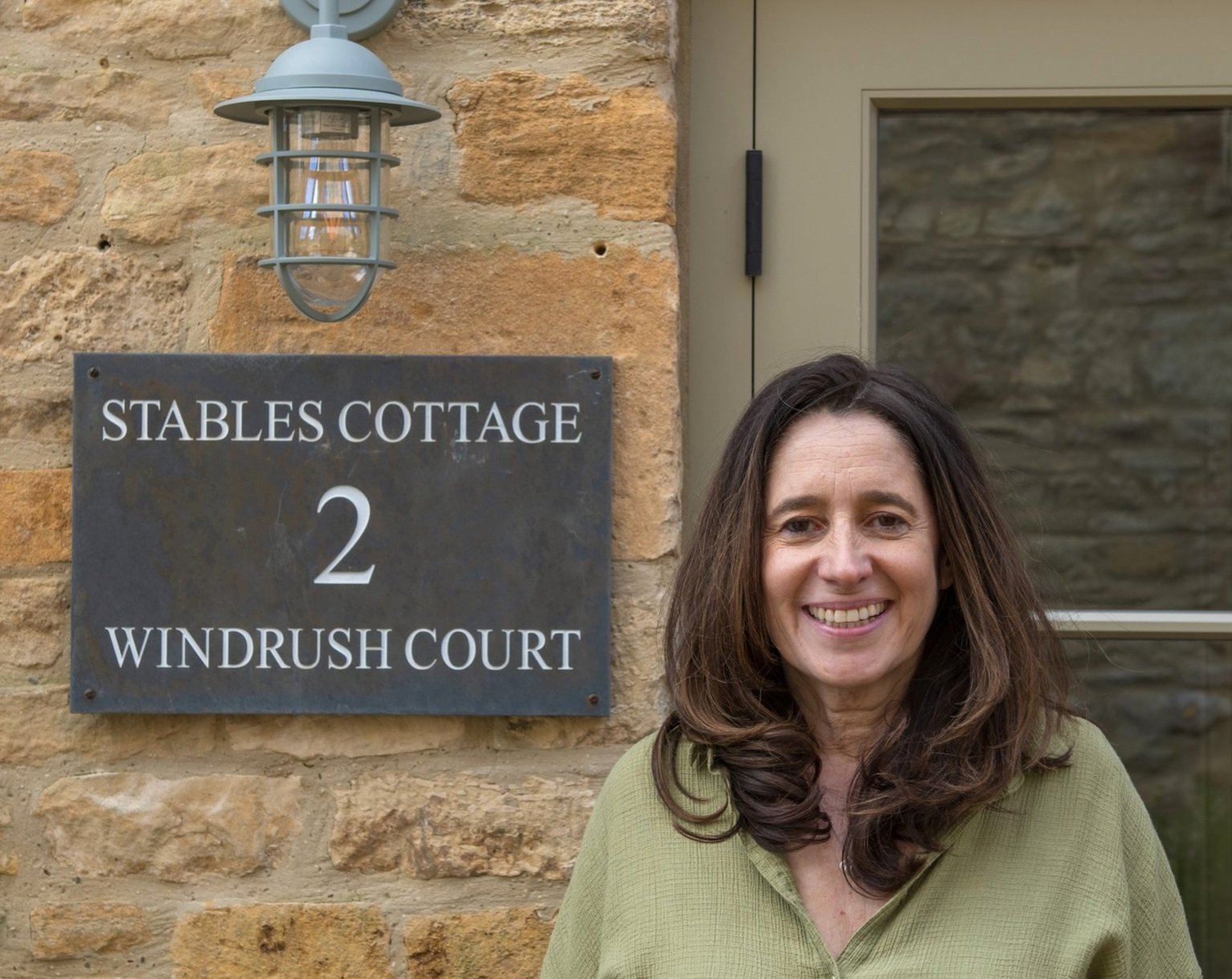 A smiling portrait of Amanda Stecker, owner of Stables Cottage, positioned next to a sign for 2 Windrush Court, located in the historic town of Stow-on-the-Wold