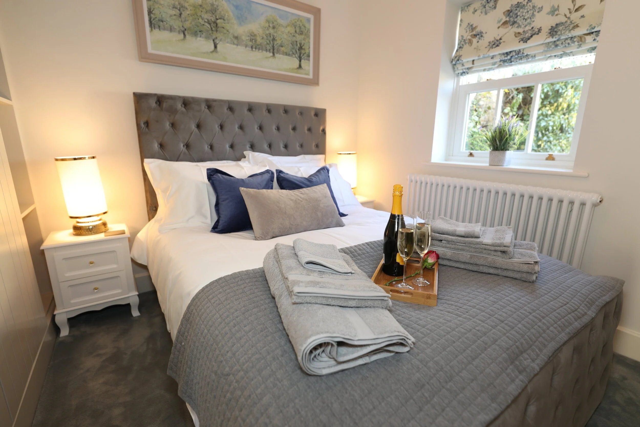 A beautifully decorated, inviting bedroom in a holiday cottage in Chipping Campden, featuring a comfortable bed with plush pillows and a welcome tray with champagne