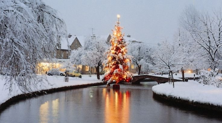 A magical snowy evening scene featuring a glowing Christmas tree reflected in the River Windrush in Bourton-on-the-Water