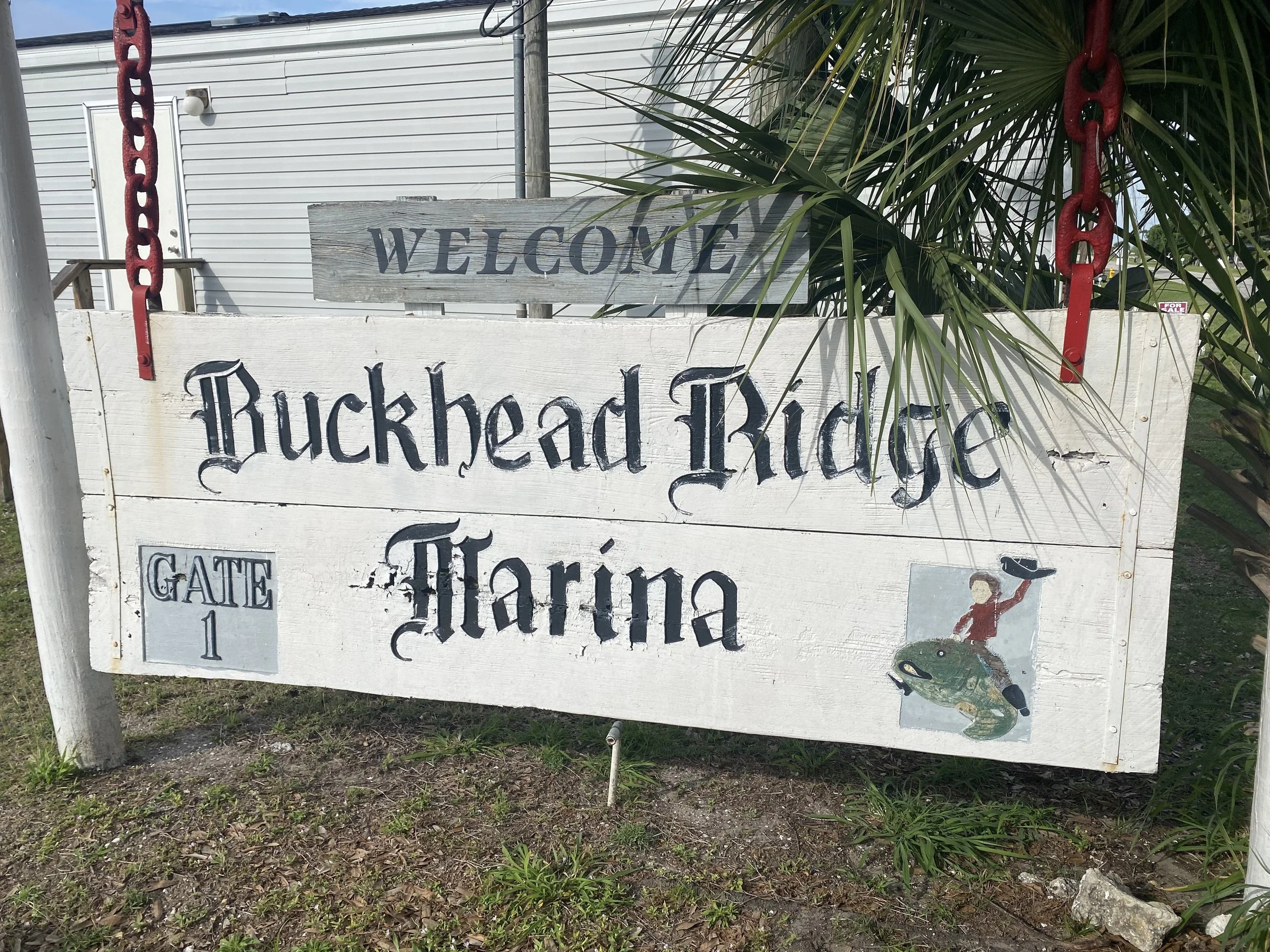 A wooden sign welcoming visitors to Buckhead Ridge Marina, indicating Gate 1, with decorative chains, a small cartoon drawing of a person fishing on a fish, and some leafy plants in the background.