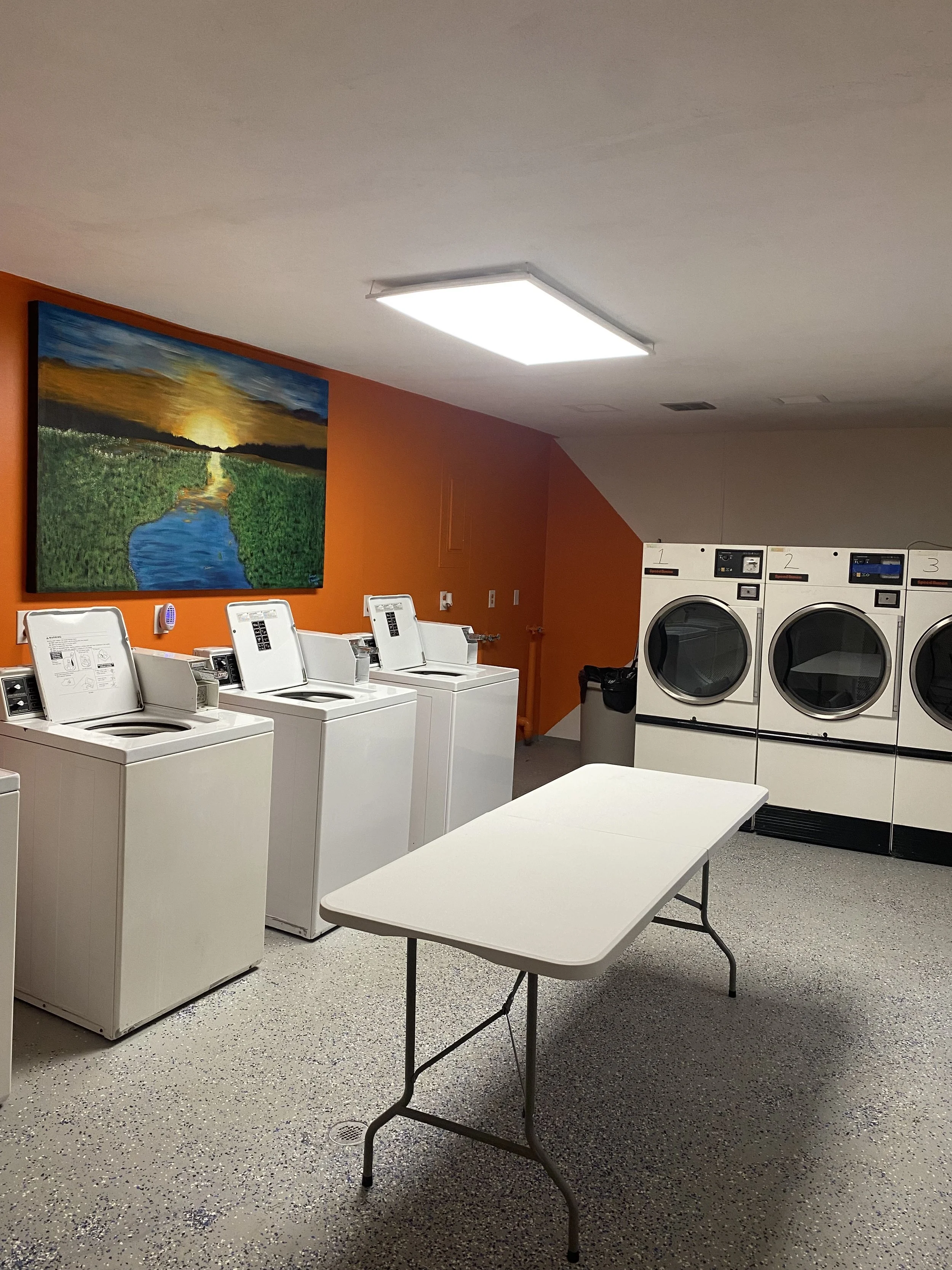A laundry room with four washing machines on the left and three dryers on the right. There is a white folding table in the center, an orange wall with a colorful landscape painting, and a ceiling light.