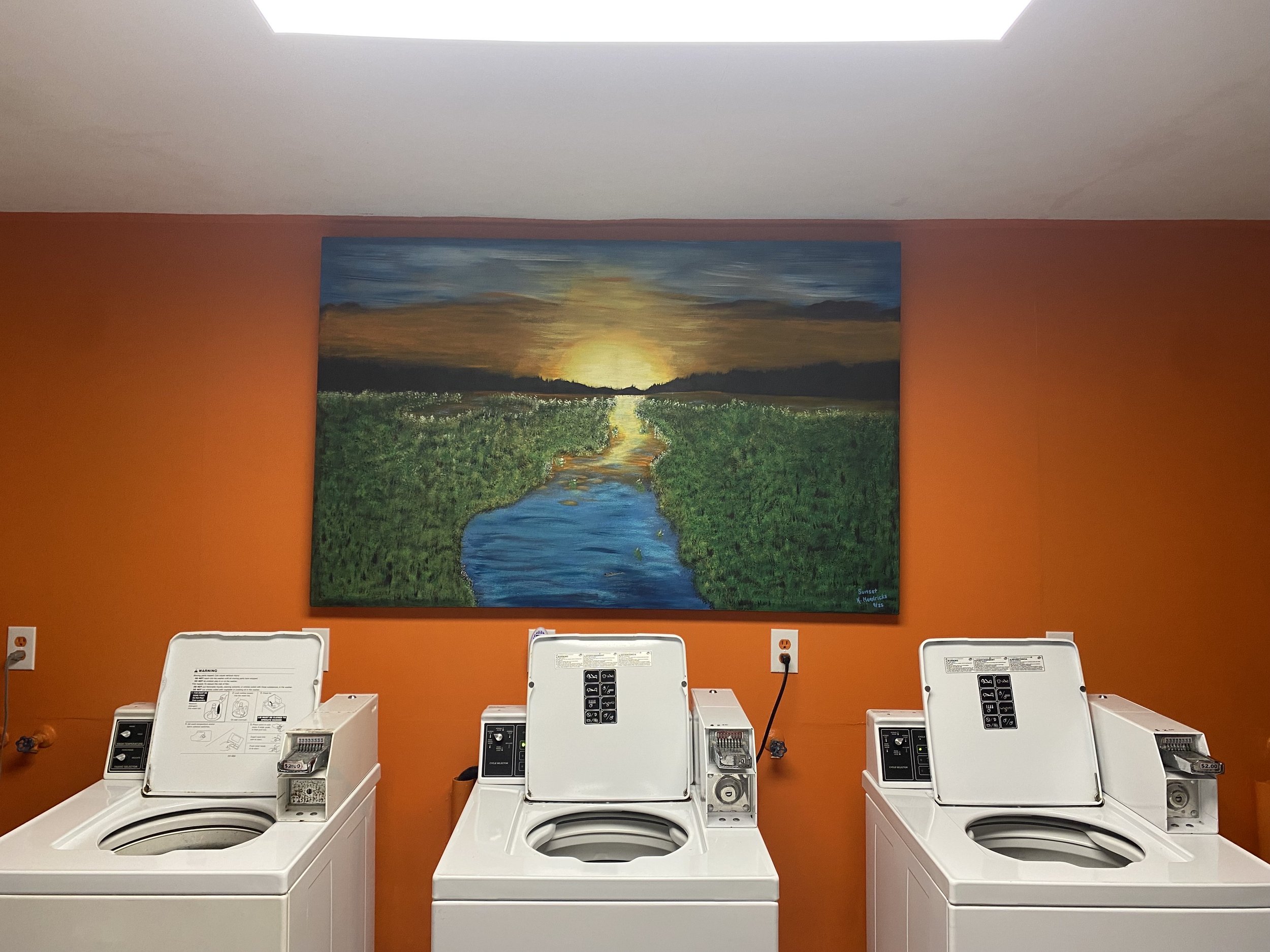 Three white washing machines in a laundromat with a colorful landscape painting of a river, green fields, and a sunset on an orange wall behind them.