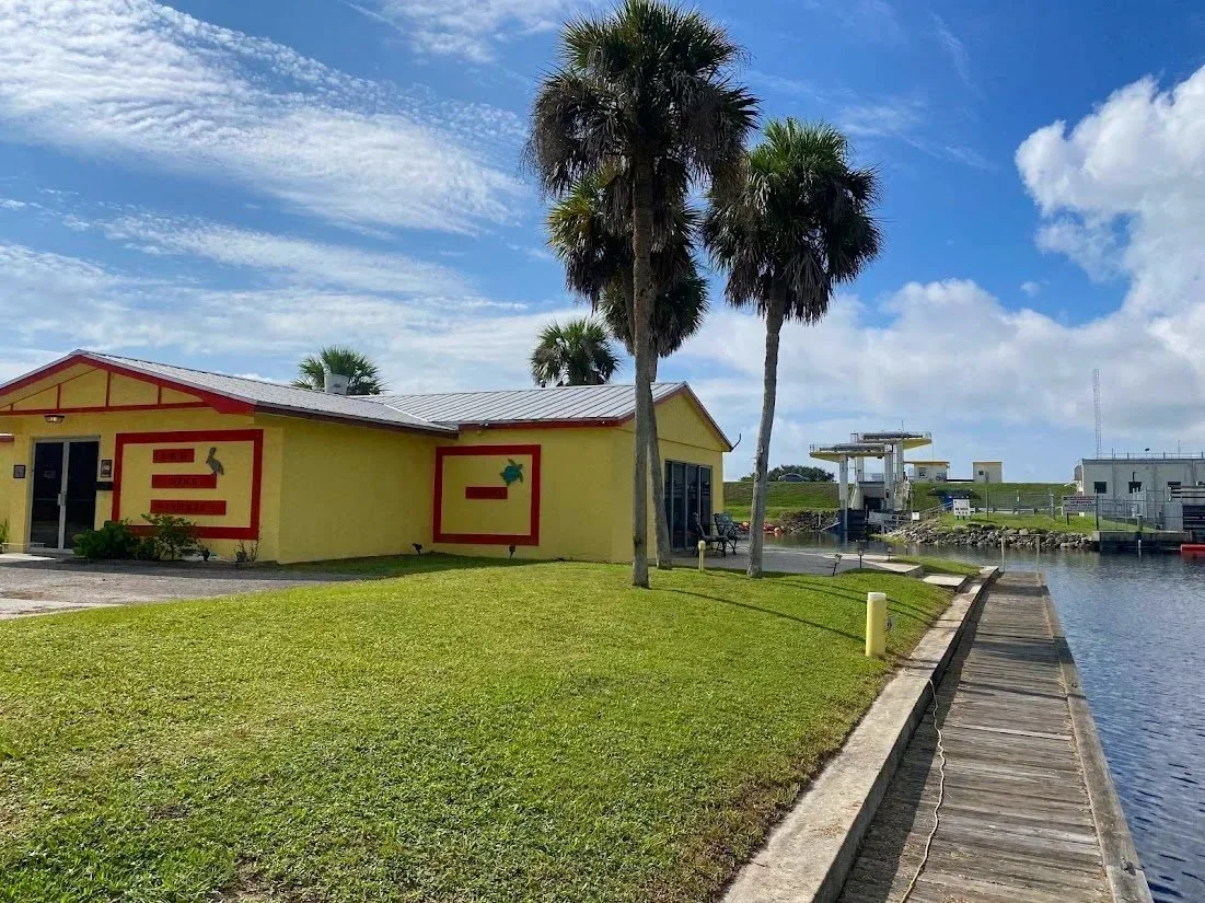 Yellow building with red trim near a waterway, with three tall palm trees in front, under a partly cloudy sky.