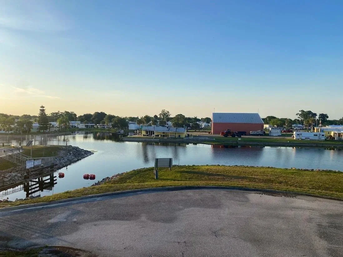 A calm lake with a small dock, surrounded by trees, buildings, and RVs, under a clear blue sky during sunset.