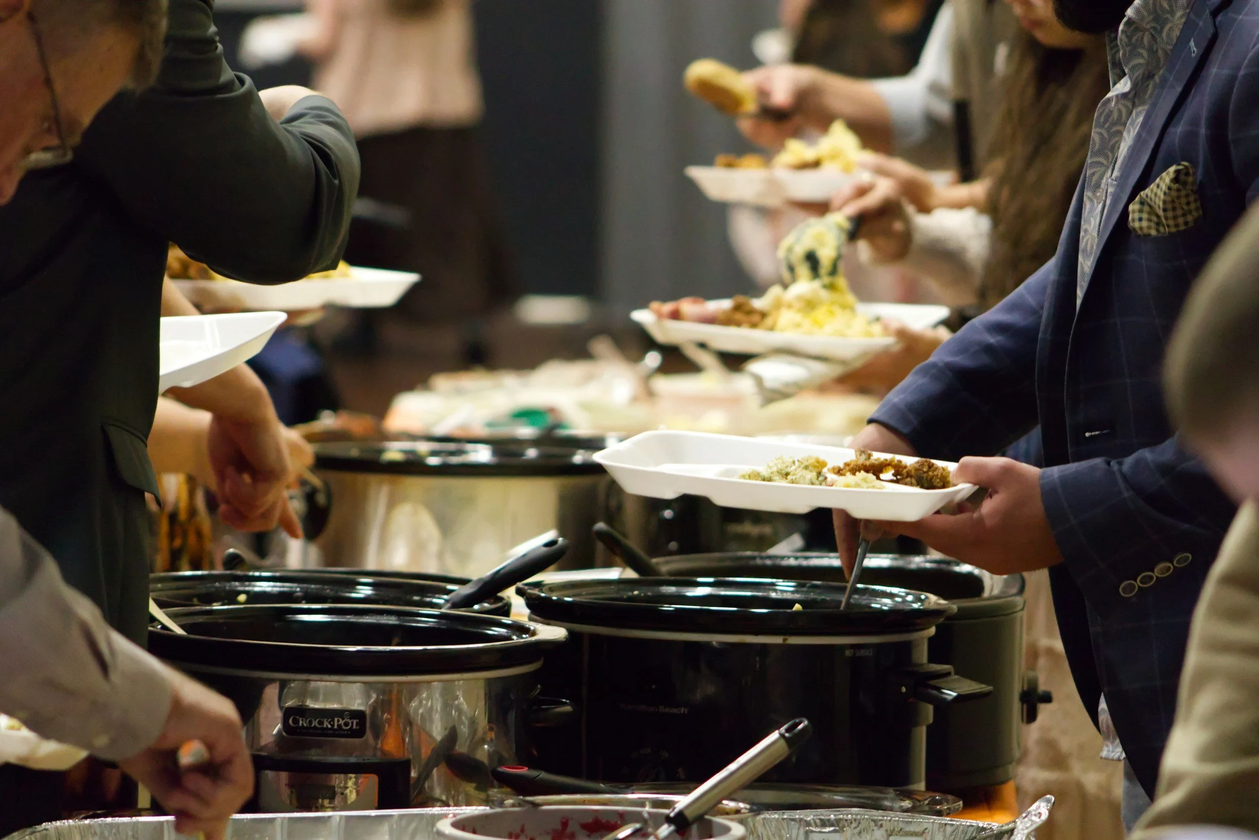 People serving themselves food at a buffet with multiple hot dishes in black slow cookers.
