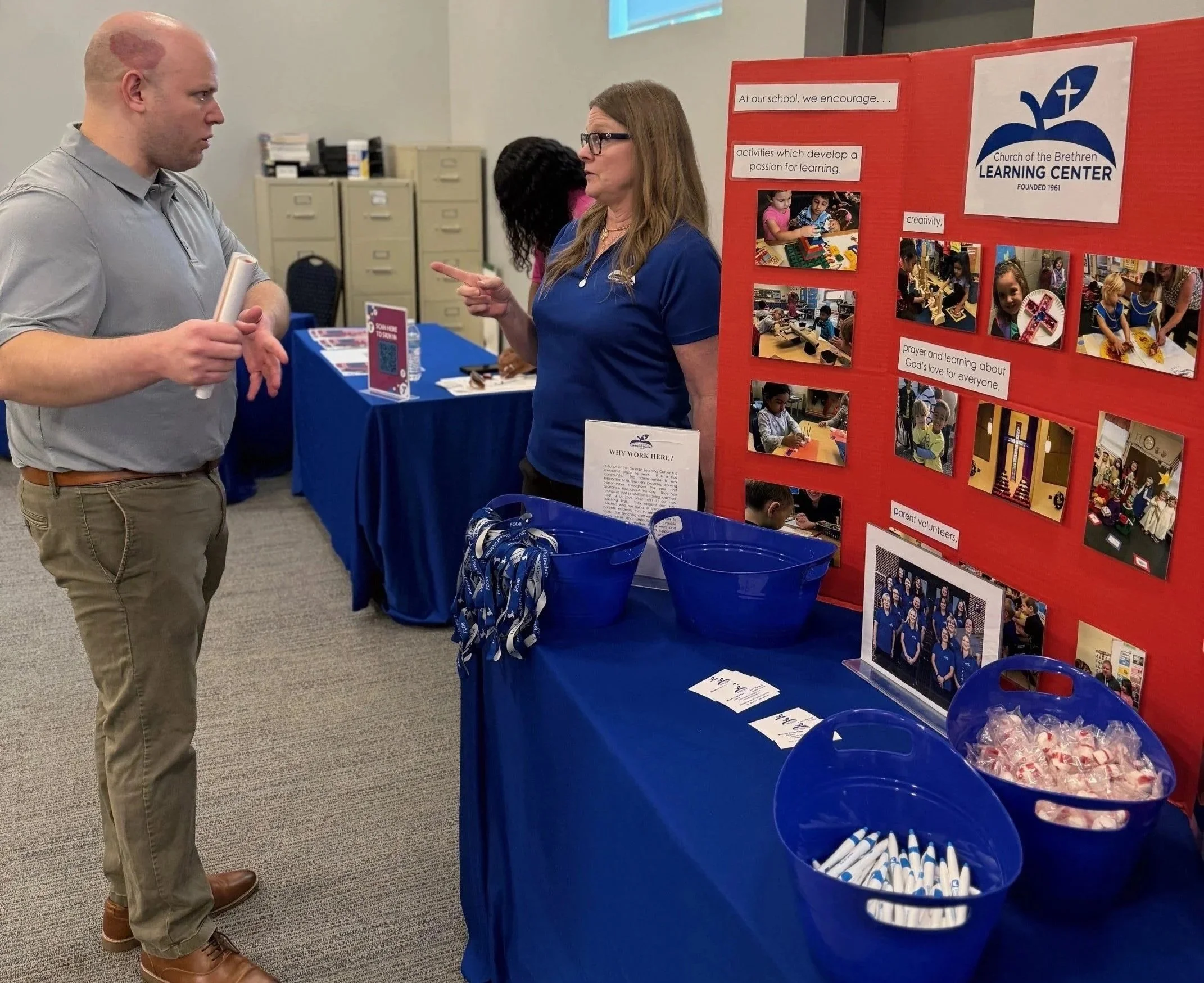 A man is talking to a women from Church of the Brethern, who is standing by a table with a red stand covered in photos showing their childcare center. Bowls with pens and mints are on a blue tablecloth.