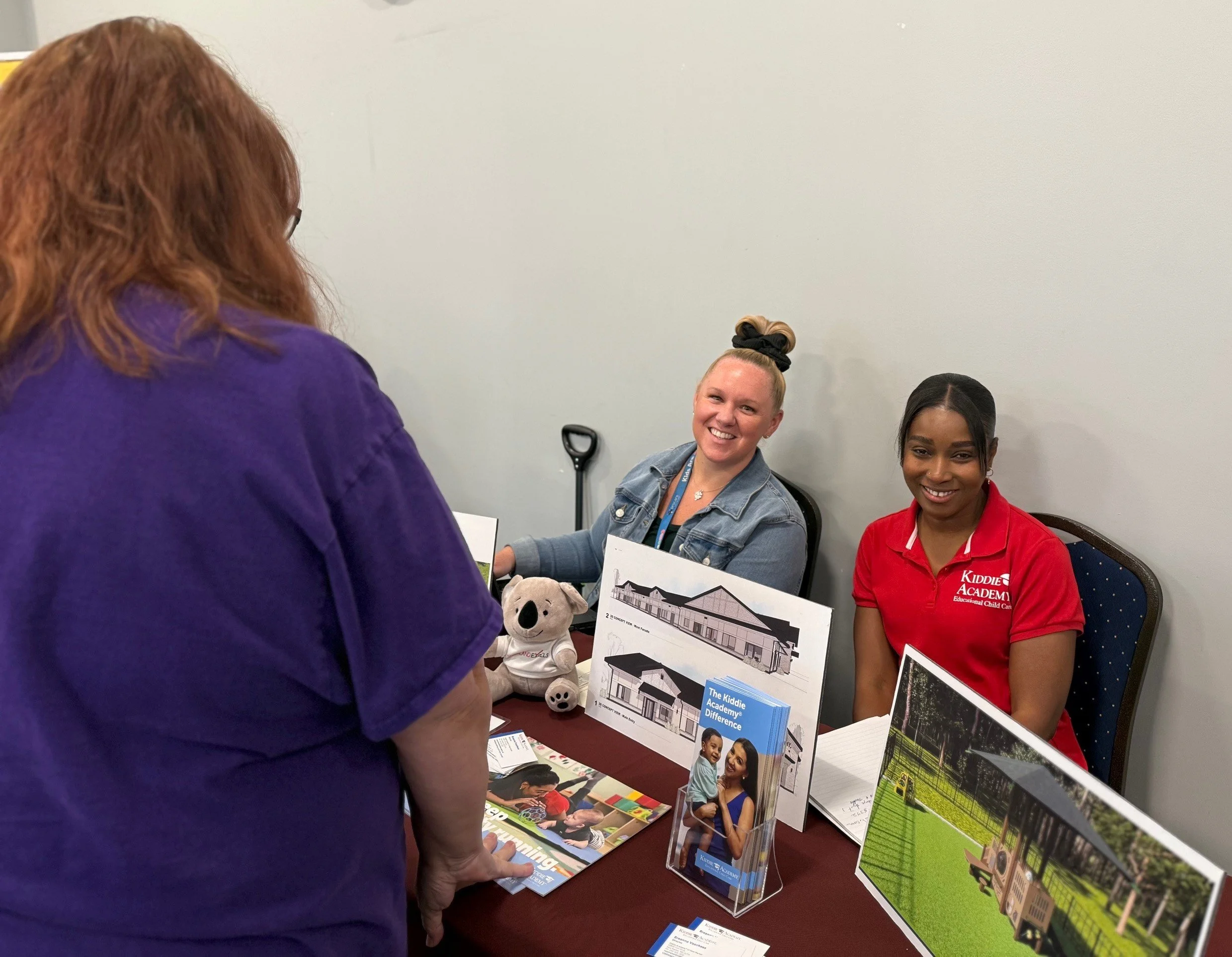 Job seeker talking to two women from Kiddie Academy, seated at a table with pamphlets about their organization.