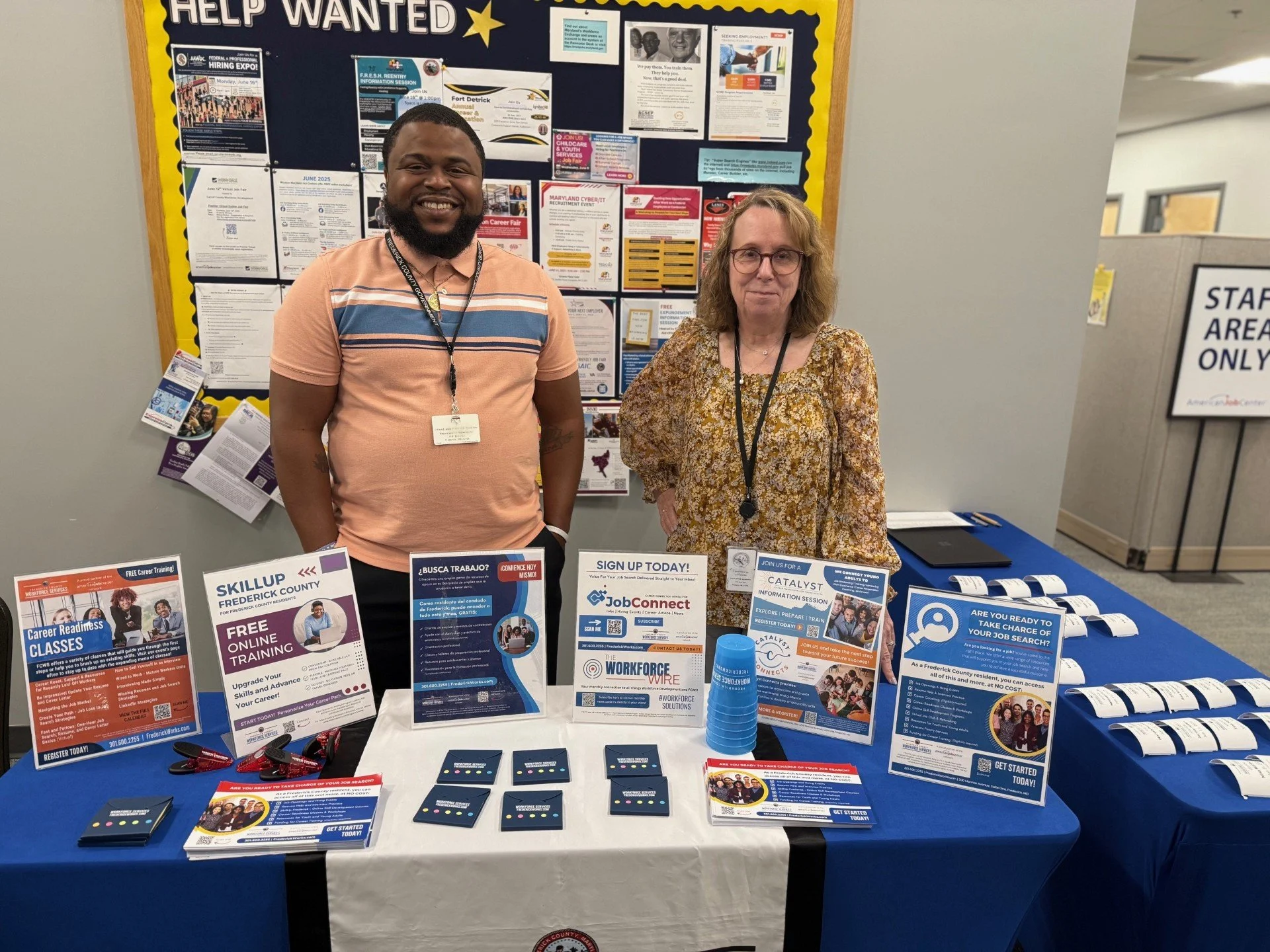 Aaron and Patti manning the Frederick County Workforce Services resource table at the Childcare & Youth Services Job Fair. Flyers describing services provided are on a table drapped with a blue tablecloth.