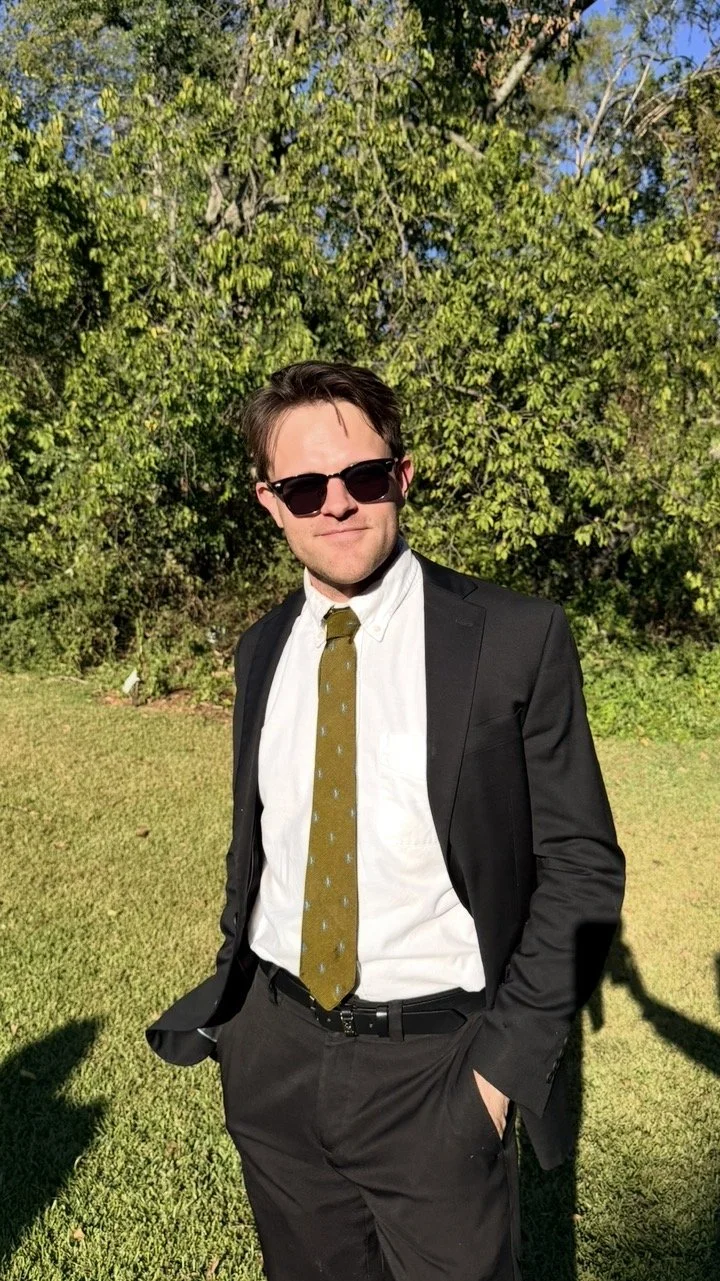 A young man in a black suit, white shirt, and green tie, wearing sunglasses, standing outdoors on grass with trees in the background.