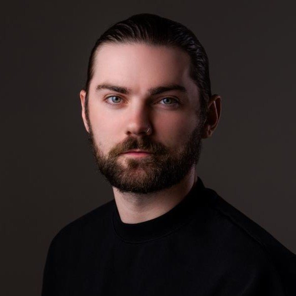 A portrait of a man with dark hair, a beard, and blue eyes wearing a black shirt against a dark background.