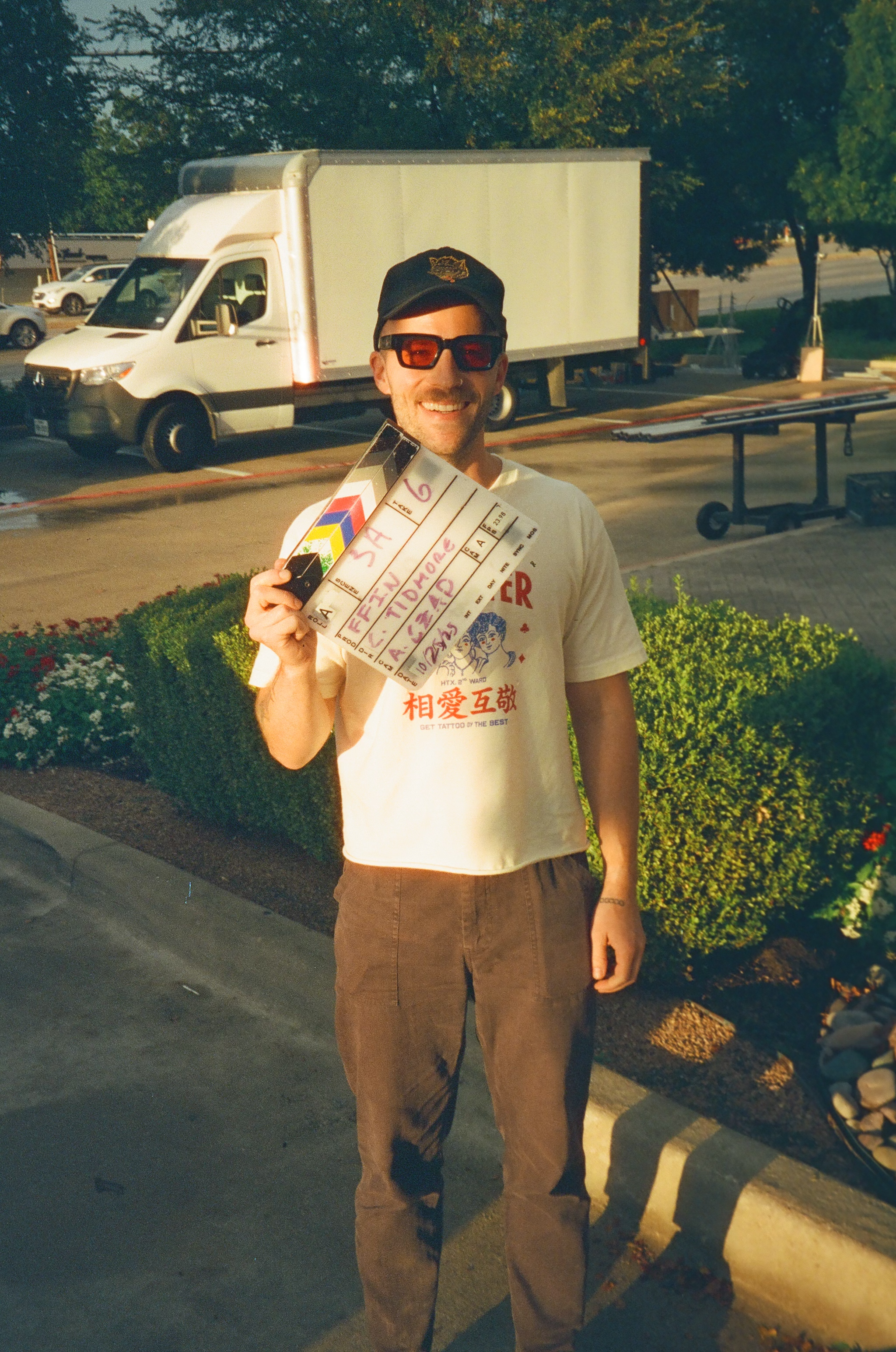 A man standing outdoors holding a film slate and smiling, with vehicles and greenery in the background.