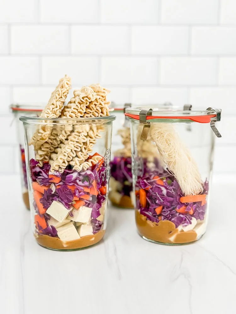 Four glass jars of meal prep for instant noodle soup containing a miso flavor base, tofu, vegetables, and noodles.